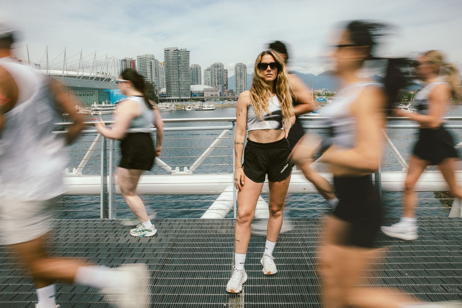 Woman in athletic wear stands still amid blurred runners on urban waterfront bridge.