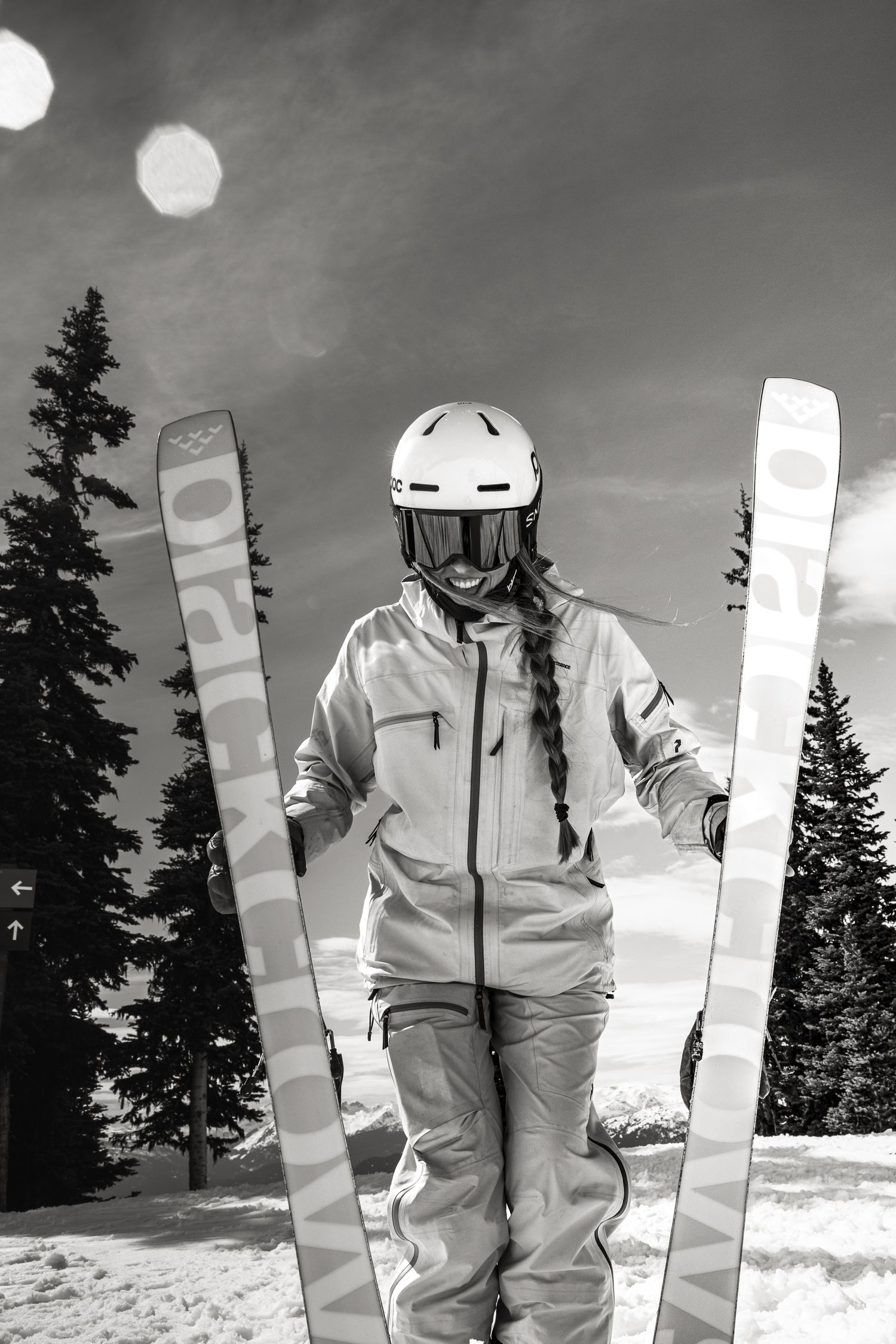 Black and white photo of female skier in white helmet holding Black Crows skis surrounded by snow-covered pine trees.
