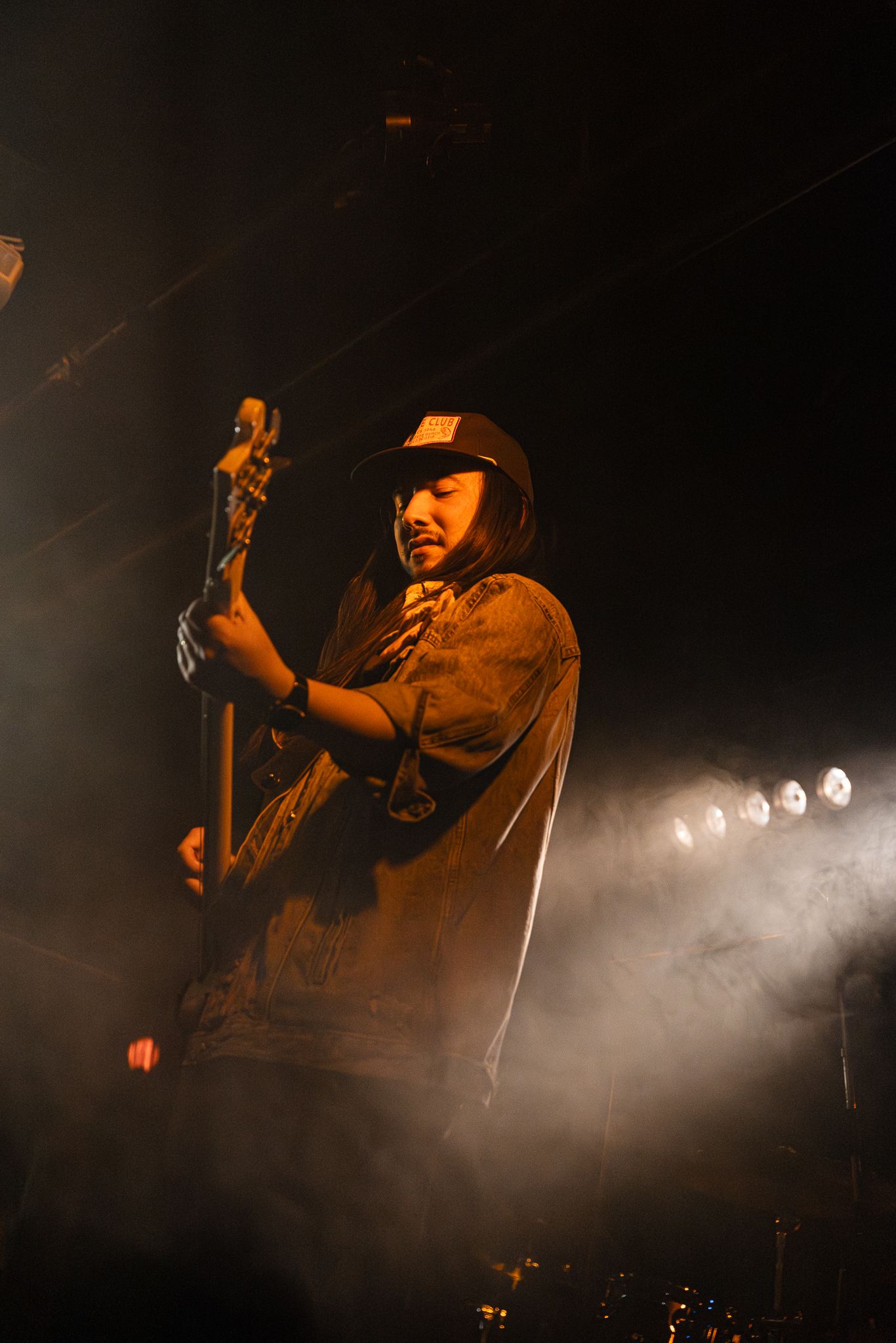 Bassist in a cap plays guitar on stage under warm orange spotlights with smoky atmospheric lighting.