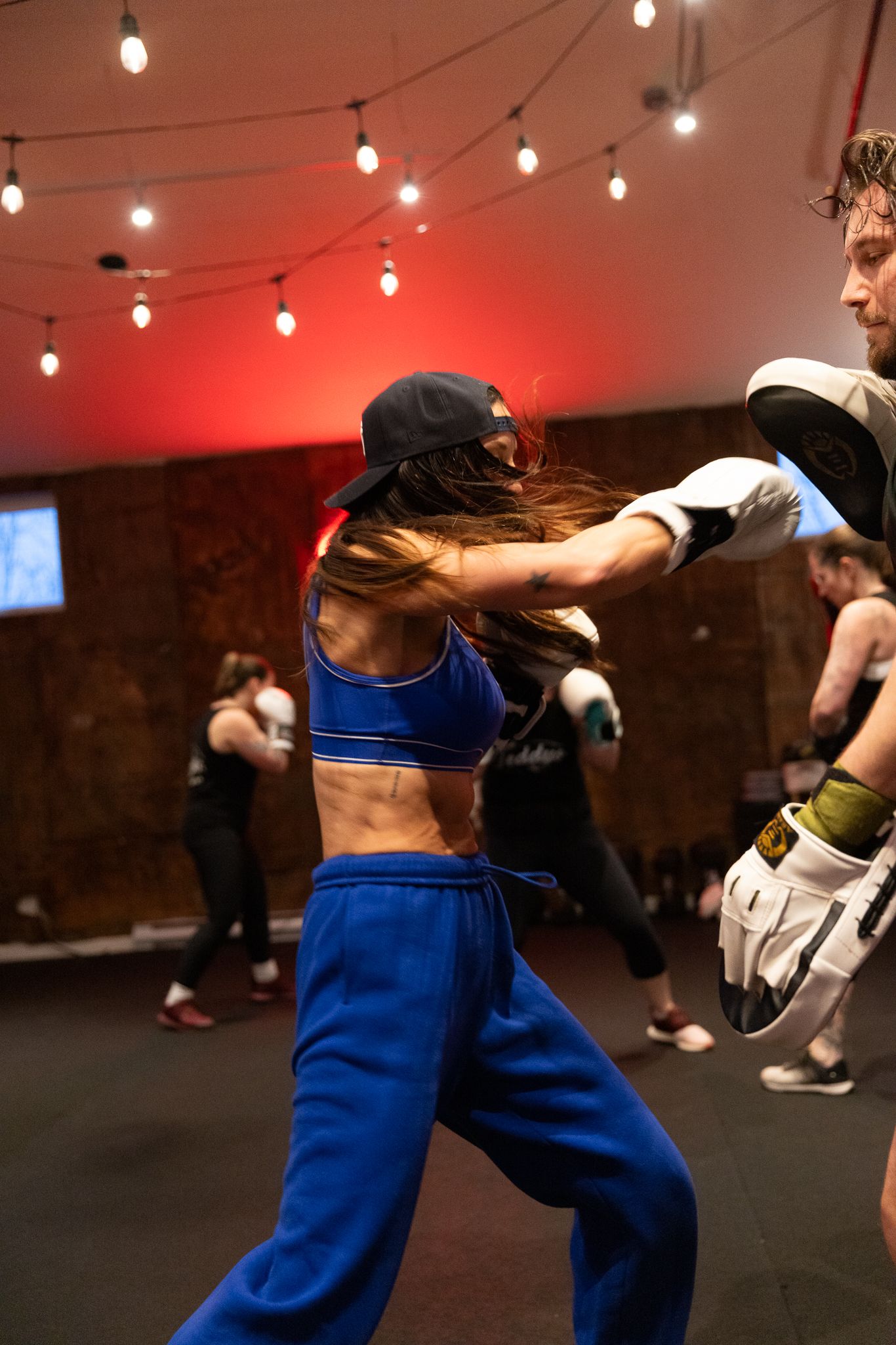 Athletic woman in blue sweats and sports bra throwing a punch at a boxing pad in a gym class.