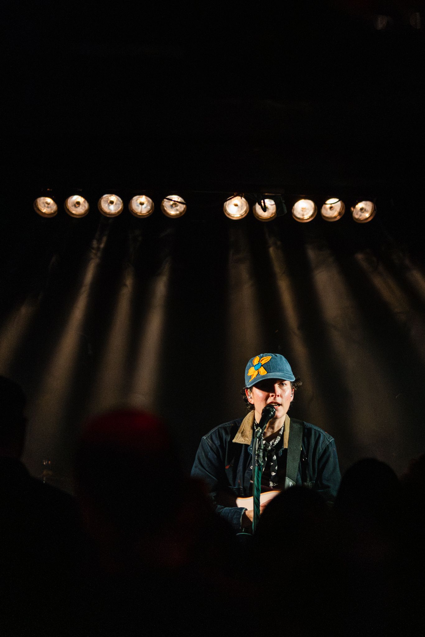 Solo performer in a star-print cap stands at a microphone beneath a row of bright stage spotlights.