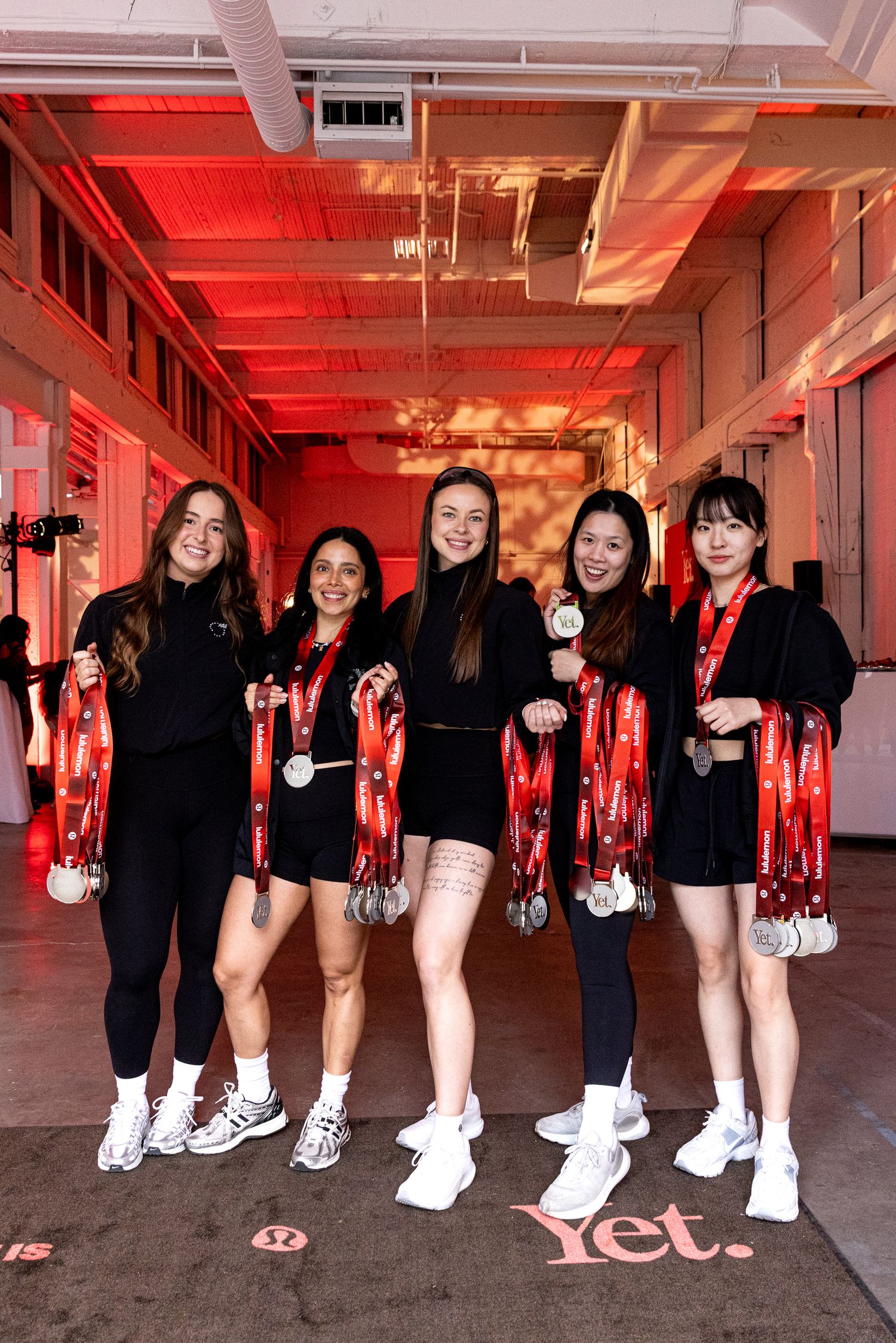 Five women in black athletic wear proudly displaying running medals at a Lululemon event.