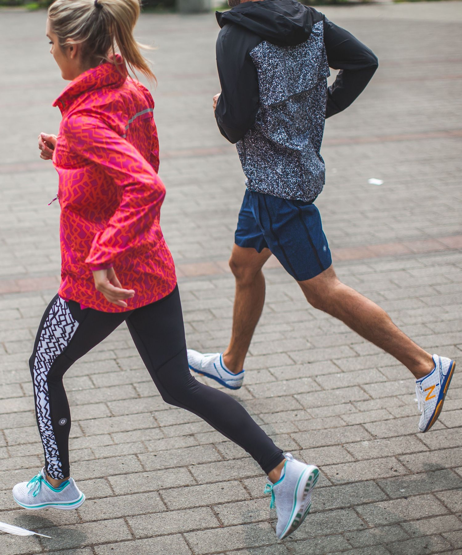 Two runners in colorful athletic gear jogging on a brick path, wearing performance running shoes and weather-resistant jackets.