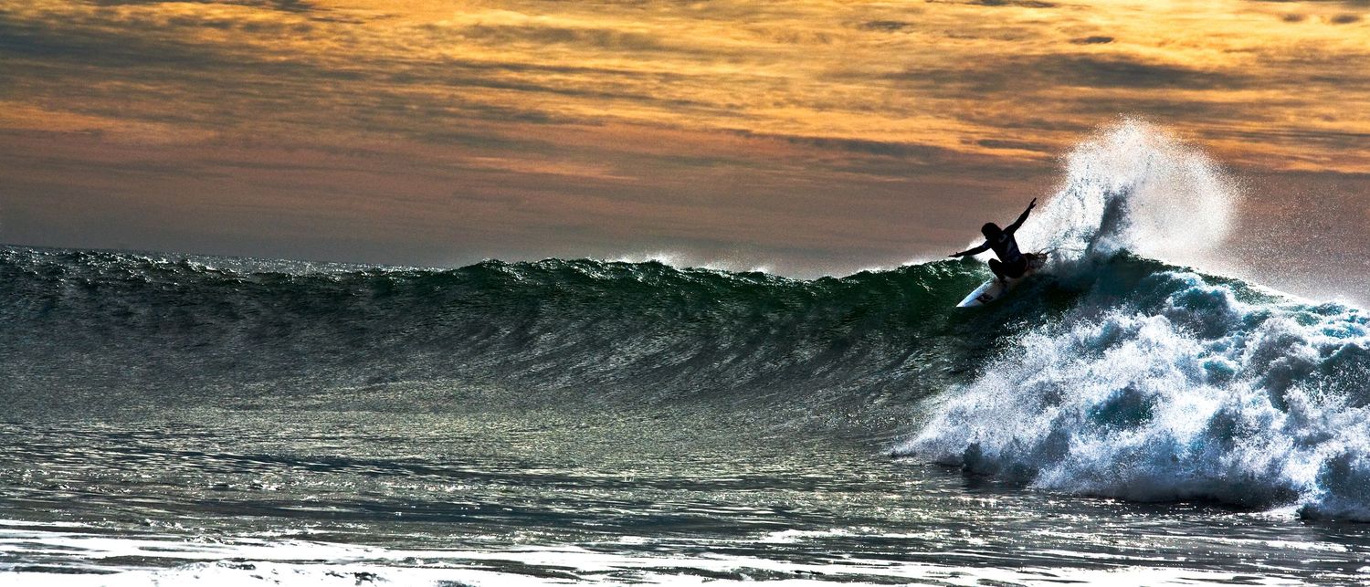 Surfer riding a massive wave at sunset with dramatic orange sky and white sea spray in the ocean.