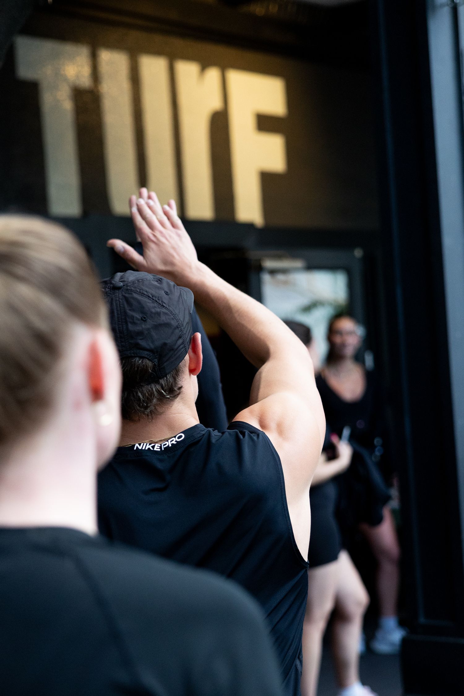 Person in Nike Pro cap raises hands outside Turf gym entrance, wearing black athletic wear.