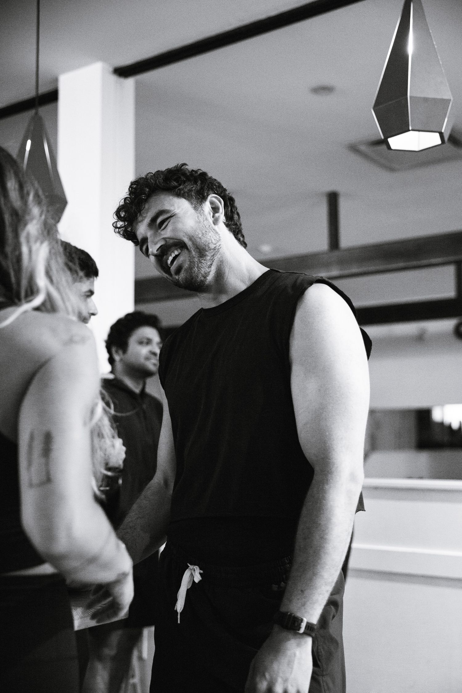 Muscular man in black sleeveless shirt laughing at a social gathering in a modern indoor space.