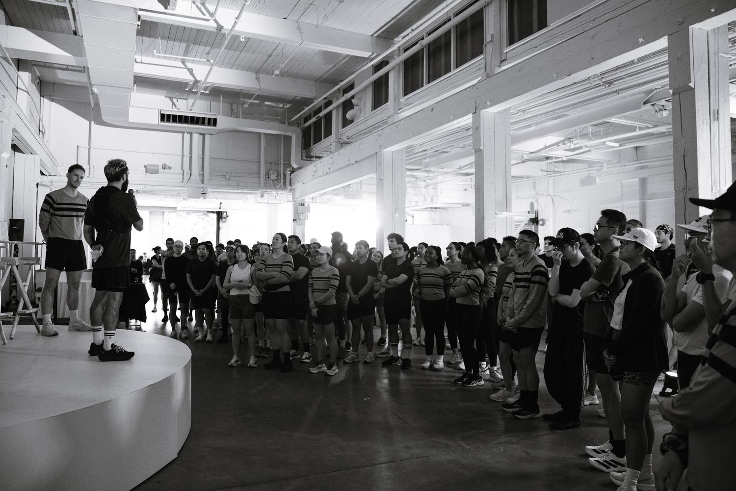 Black and white photo of a large group gathered indoors listening to a speaker on a platform in a modern venue.