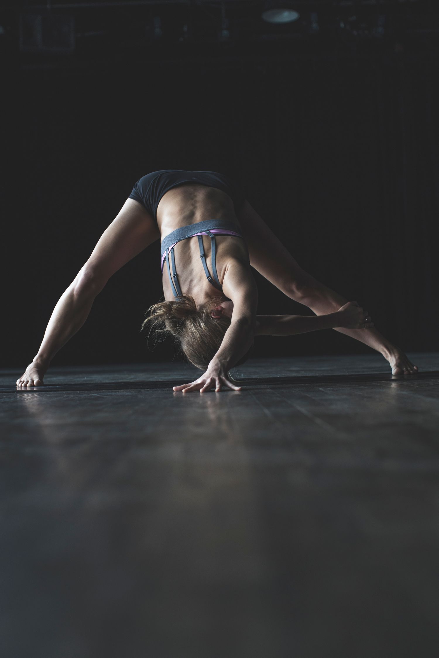 A flexible woman in sportswear performs a wide-legged forward bend on a dark studio floor.
