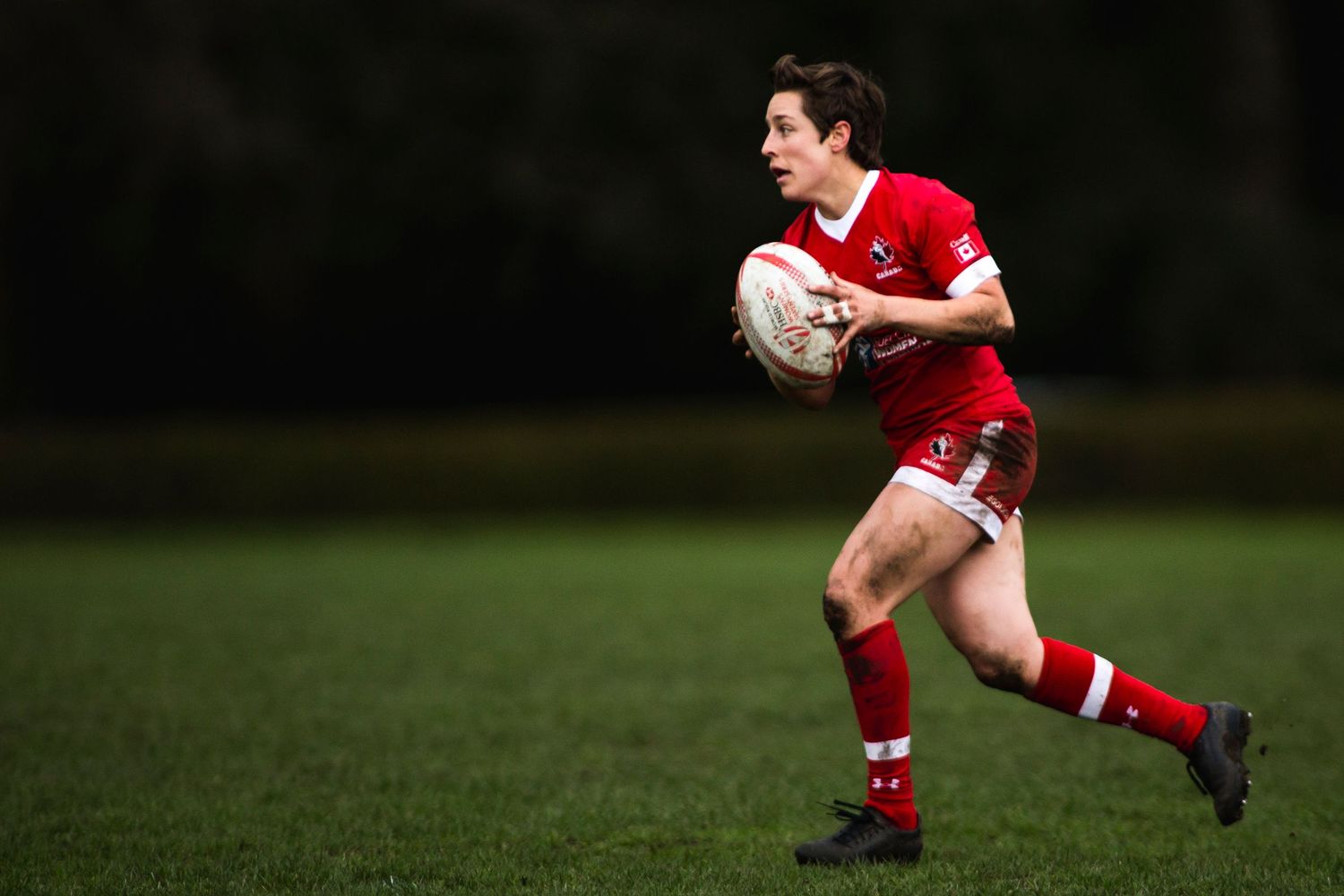 Rugby player in red kit sprinting with ball during an outdoor match on a green field.