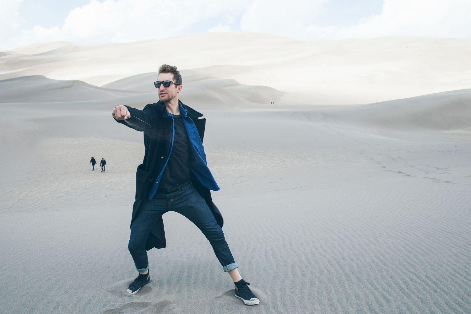 Man in black coat and sunglasses striking a pose on sandy desert dunes with two people walking in background.