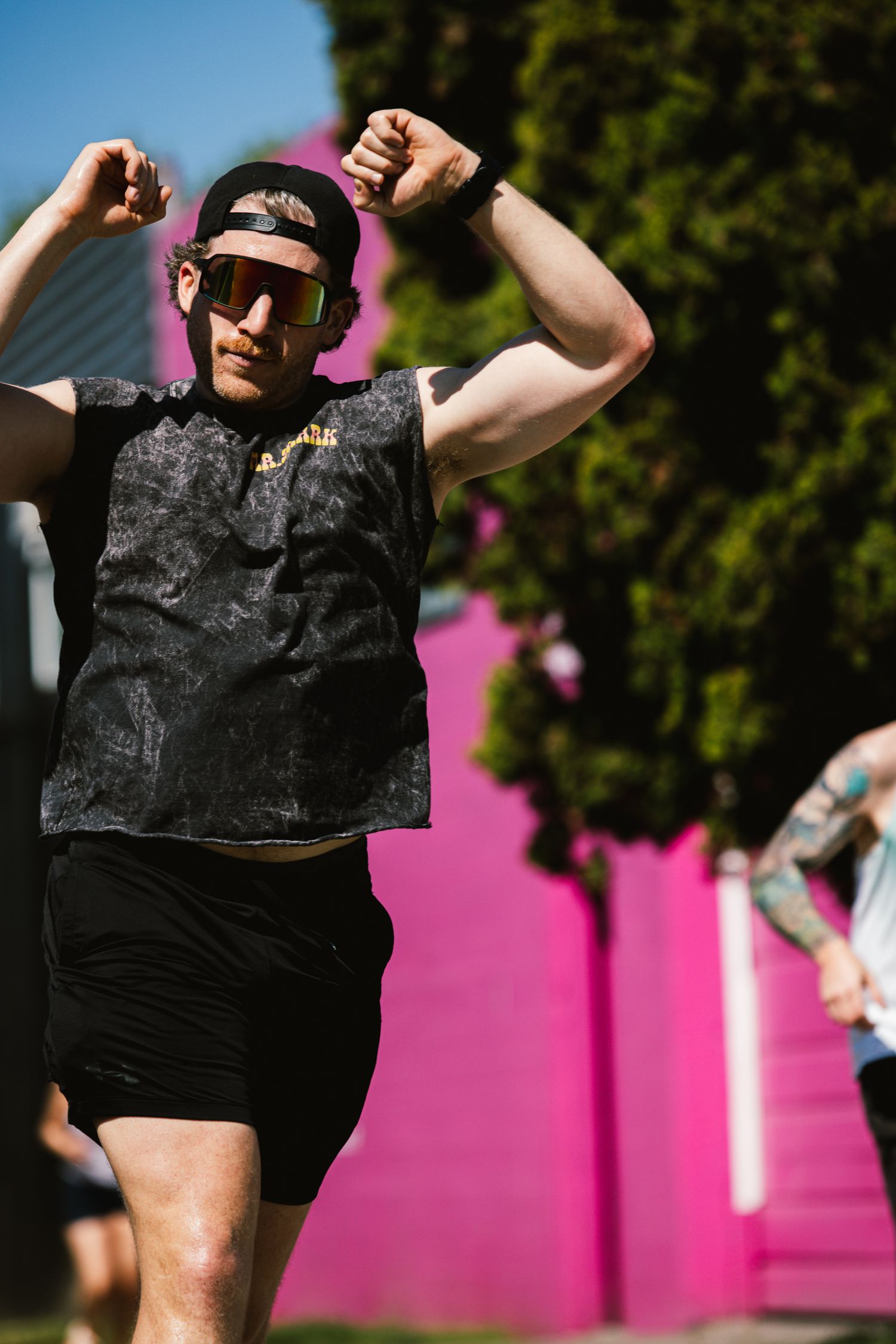 Athletic man in black crop top and cap flexes arms outdoors near a bright pink wall.