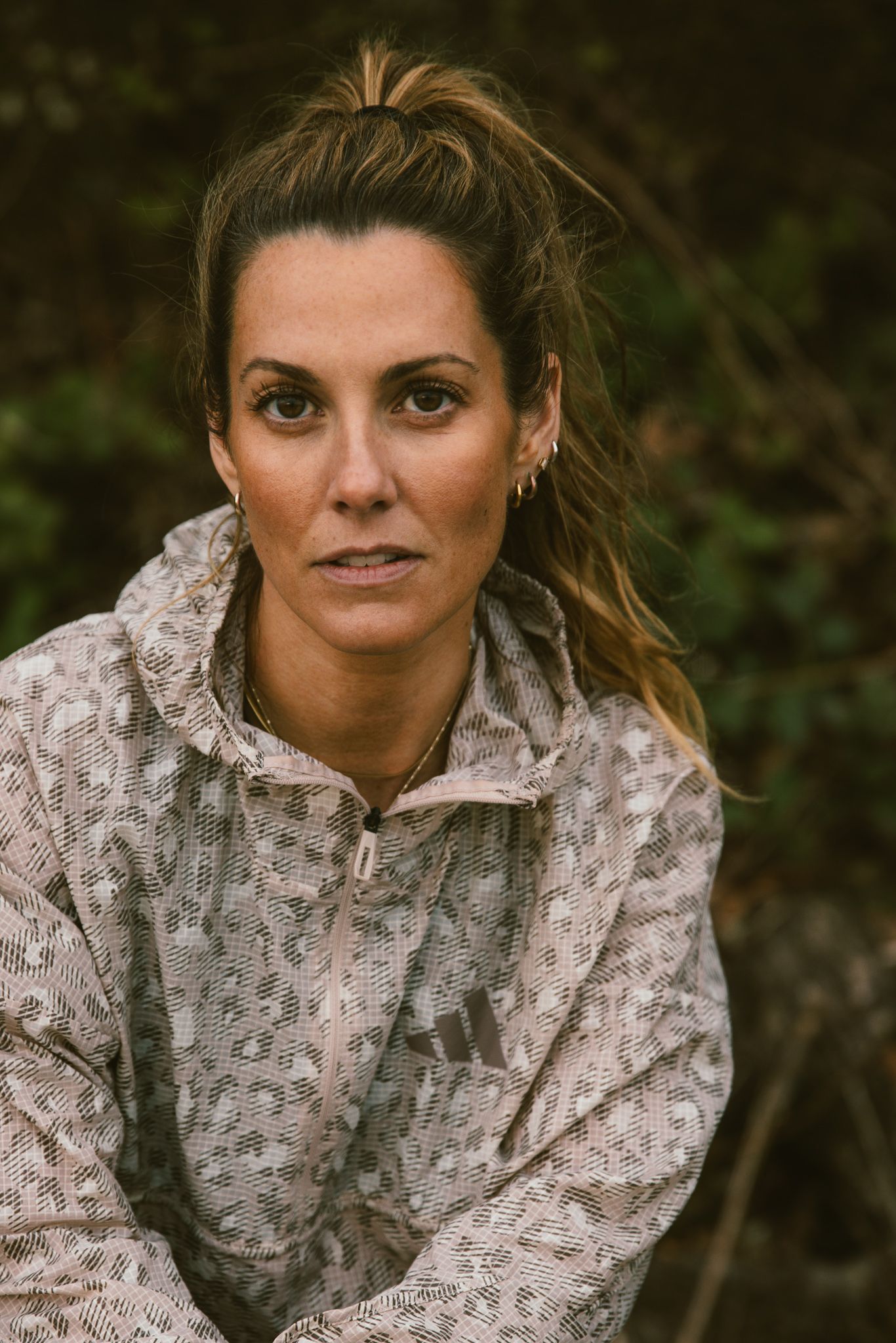 Woman in patterned Adidas athletic jacket with hair in ponytail outdoors in nature.