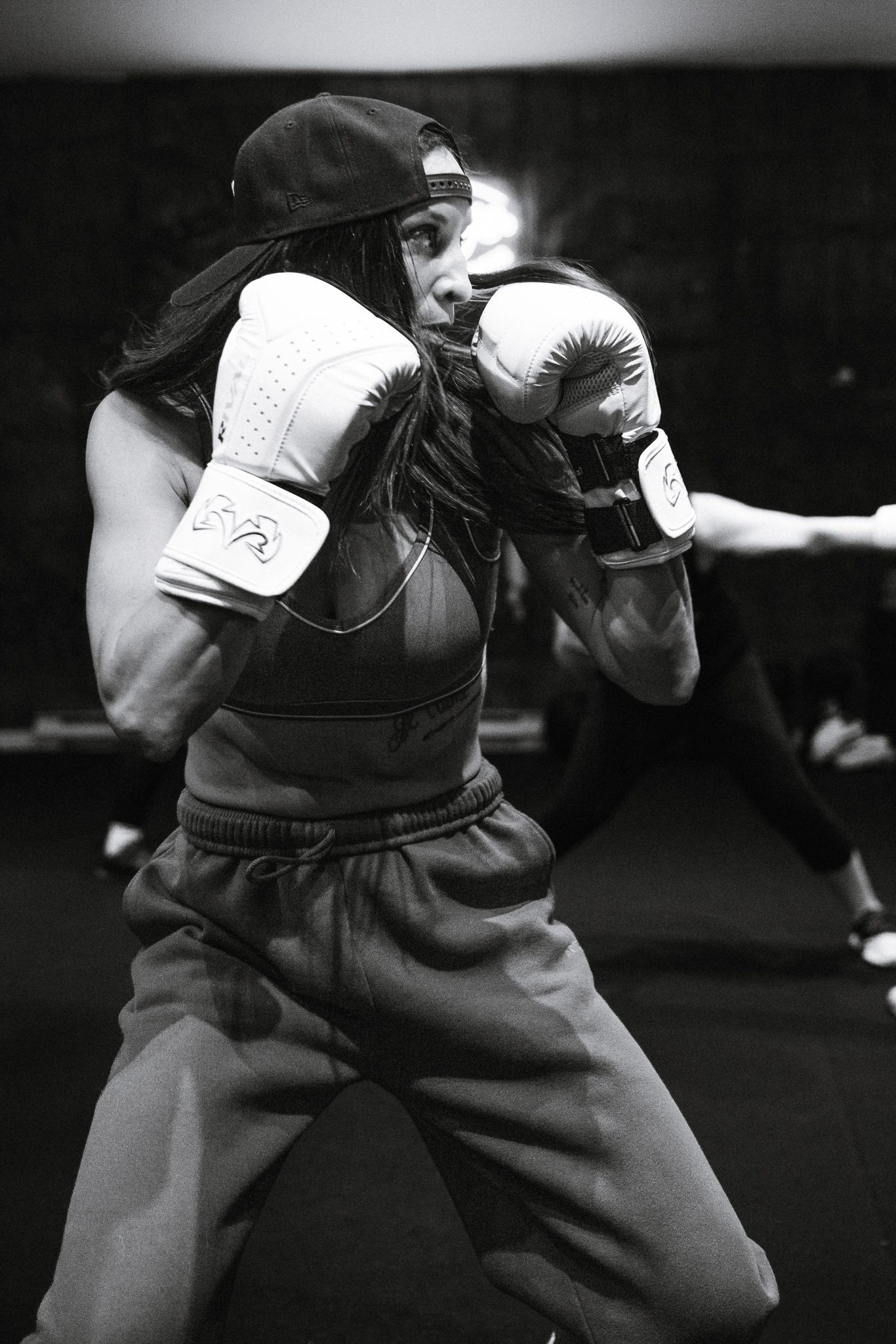 Woman in boxing gloves and cap practicing fighting stance in black and white gym photo.