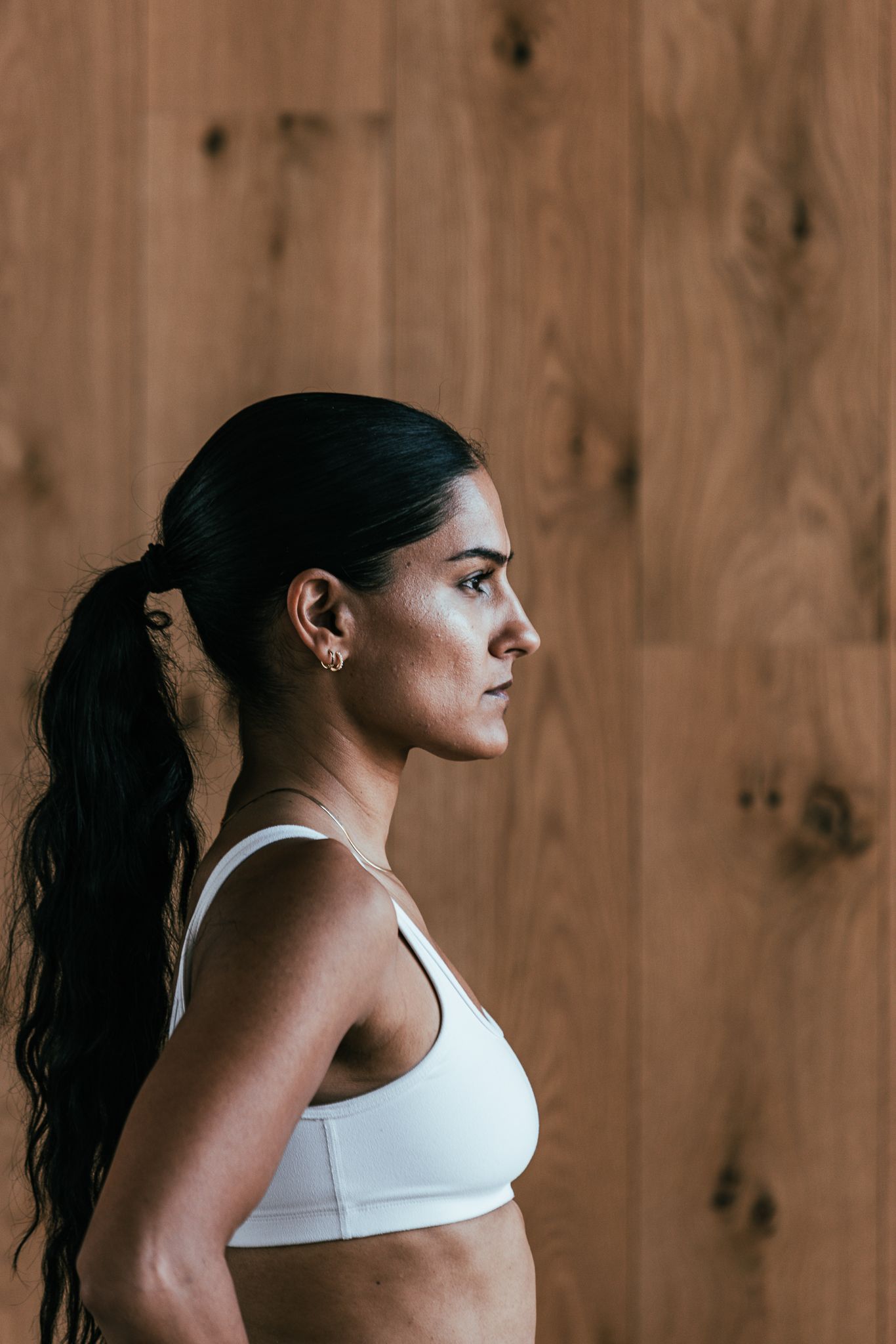 Fit woman in white sports bra with ponytail standing in profile against wooden wall background.