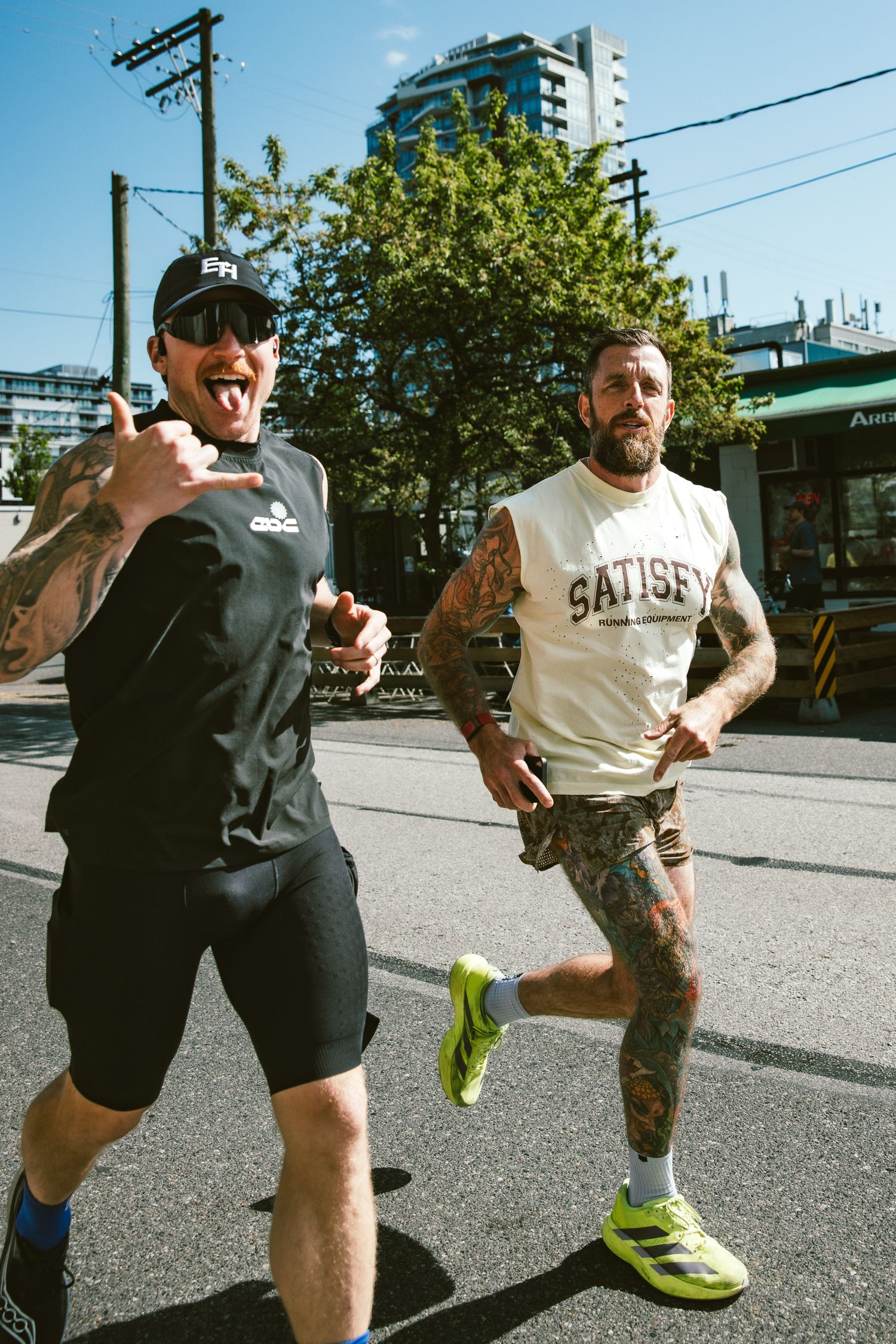 Two tattooed men in athletic wear jogging on a city street, one wearing Adidas and one in a Satisfy running vest.
