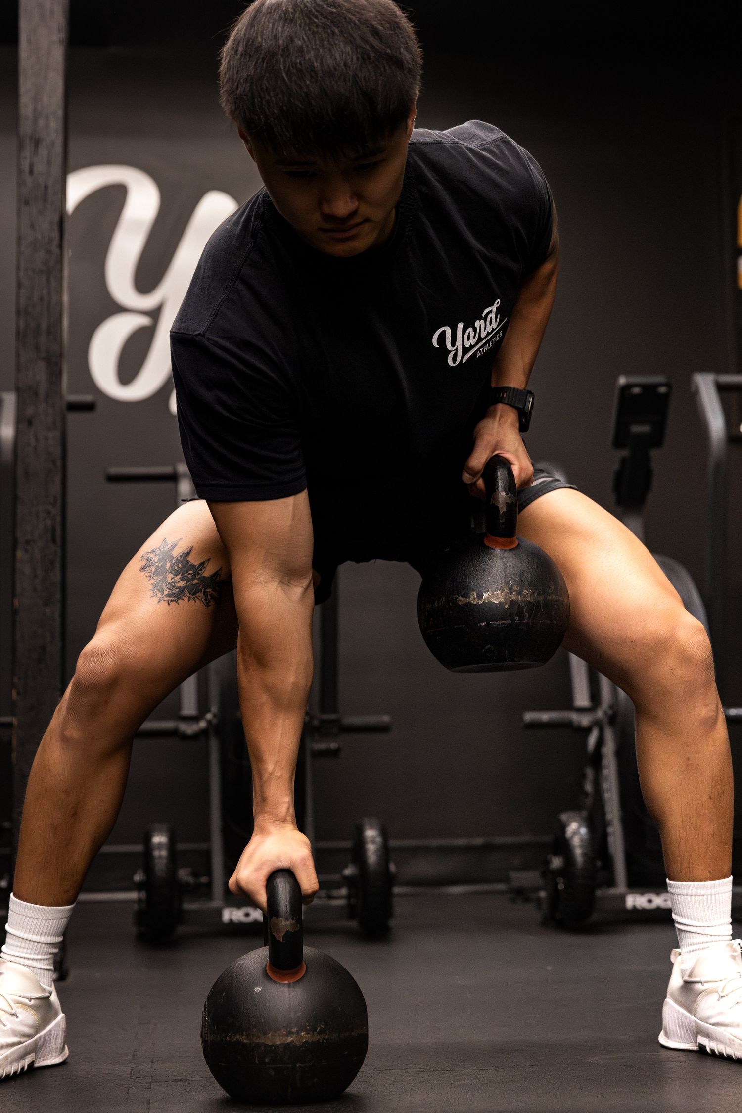 Athletic man lifting two kettlebells in a dark gym wearing black Yard brand shirt.