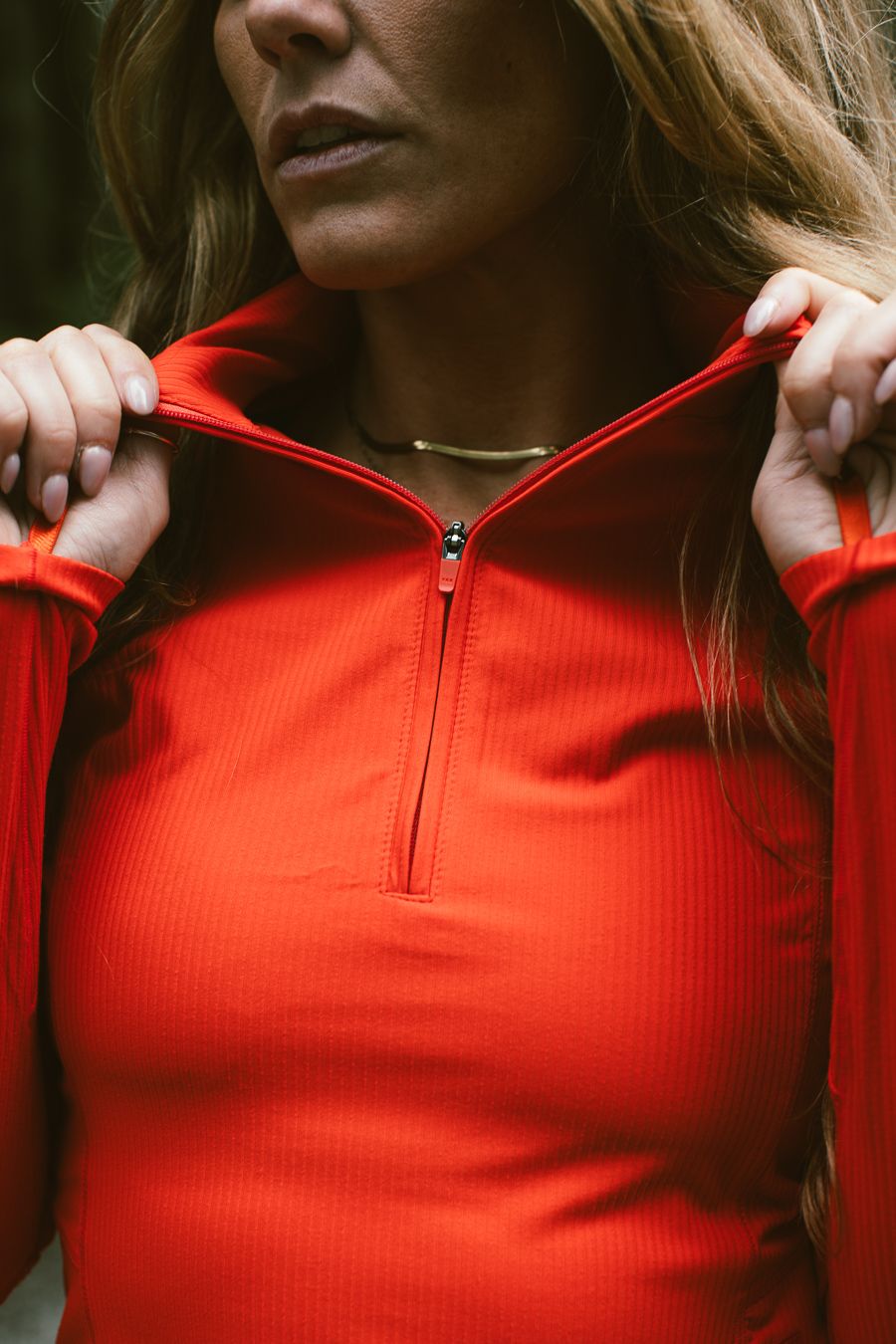 Close-up of a woman in a red quarter-zip athletic top adjusting her collar, wearing a gold necklace.