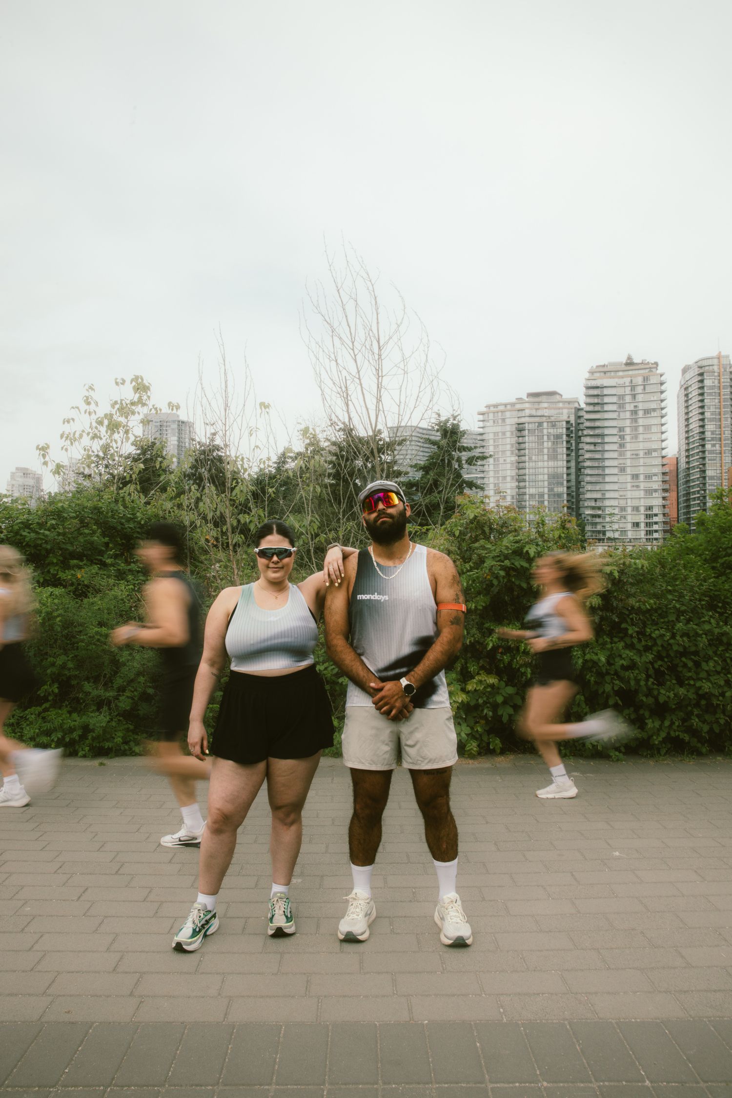 Two athletes pose together on a city running path with blurred runners passing by and urban skyline in background.