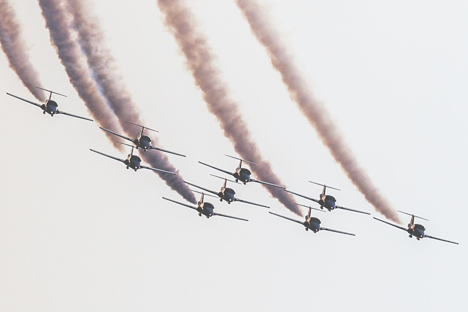 Nine military jets fly in tight diagonal formation trailing white smoke against an overcast sky at an airshow.