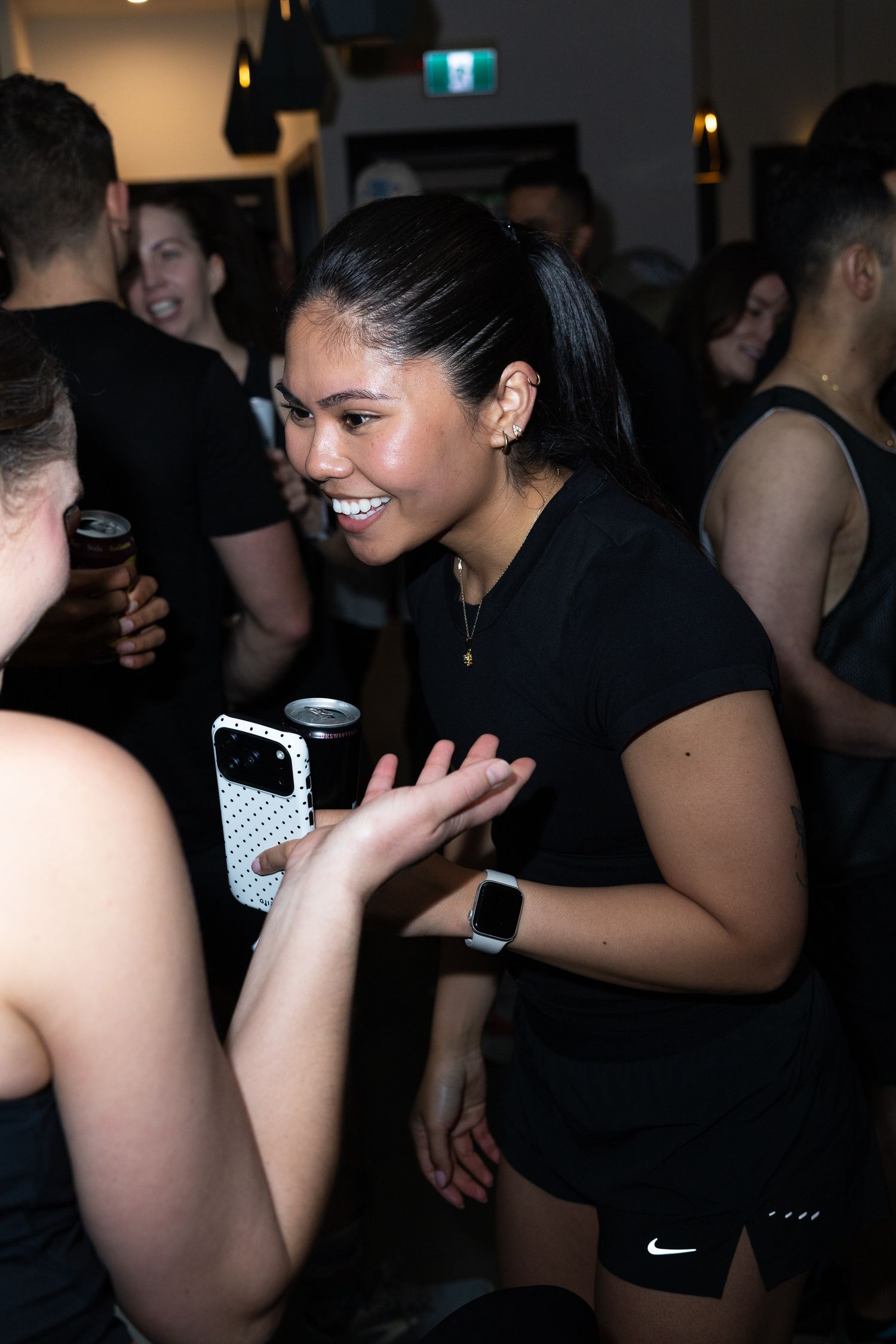 Young woman in black Nike outfit smiles while socializing at a fitness social event indoors.