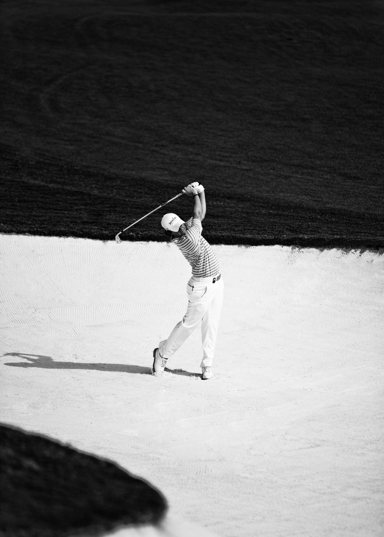 Black and white photo of a golfer completing a swing shot from a sand bunker on a golf course.