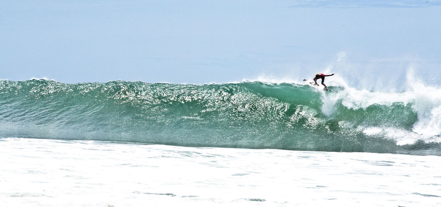 Surfer riding a large ocean wave at a sunny beach break with white water foam in the foreground.