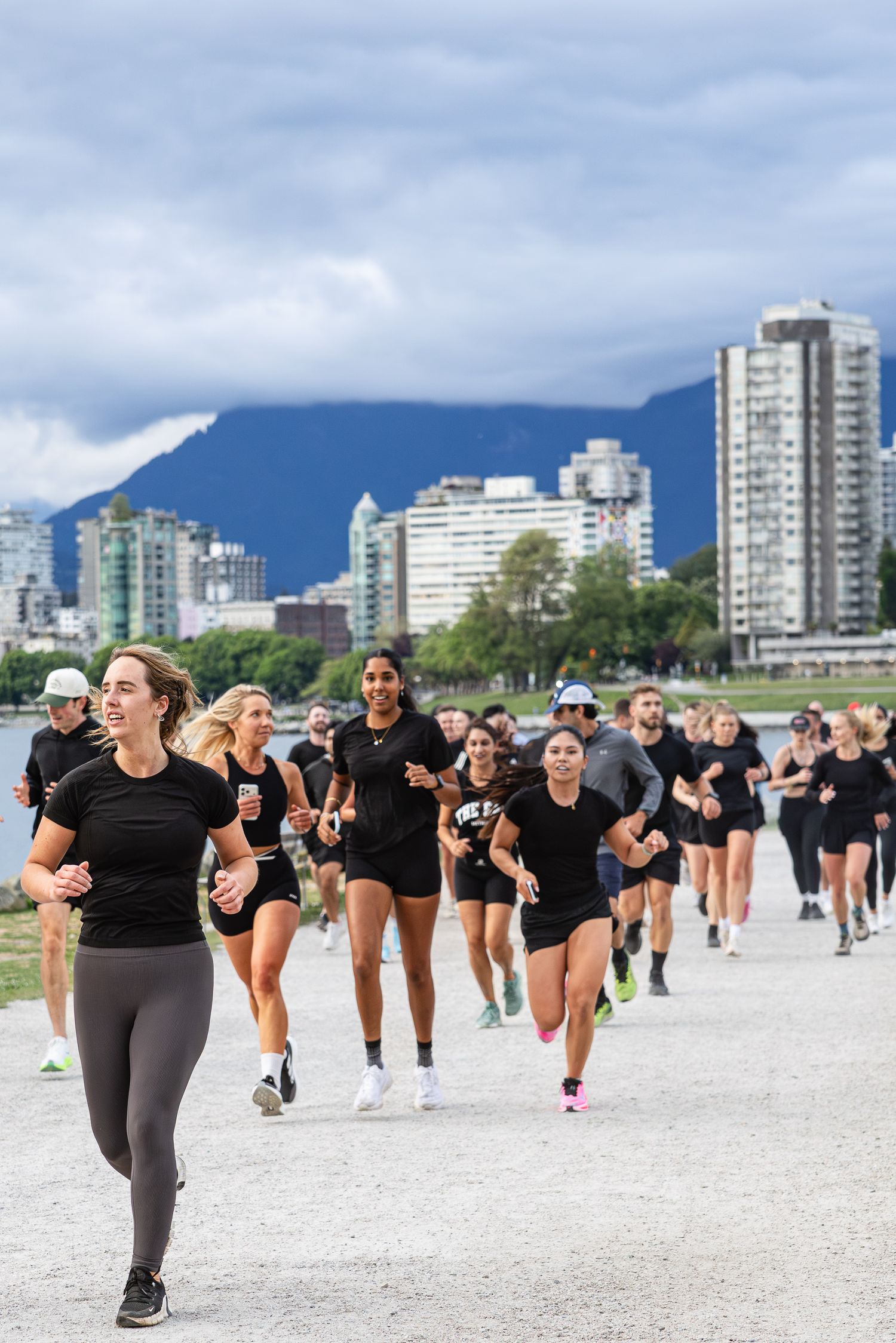 Large group of runners in black athletic wear jogging along a waterfront path with city skyline.