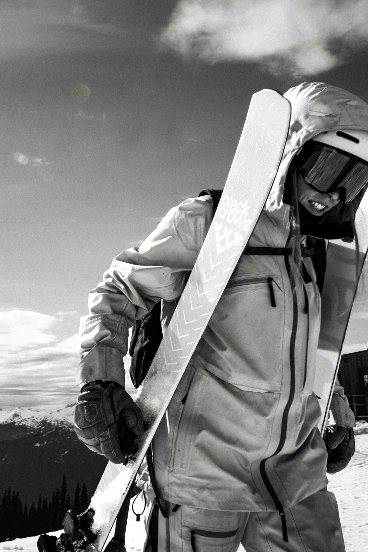 Black and white image of smiling female skier carrying skis over shoulder wearing white helmet against bright sky.