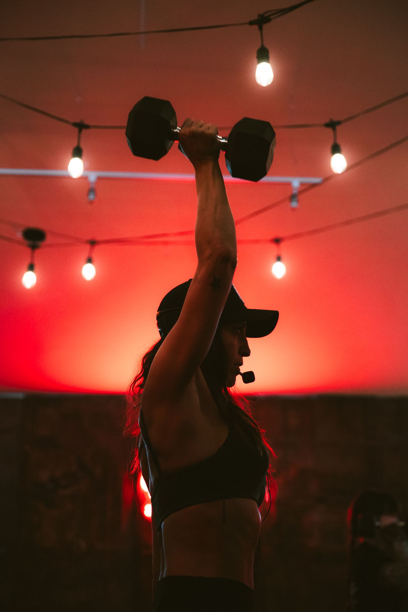 Fit woman in sports bra lifting a dumbbell overhead in a gym with warm string lights and red background.