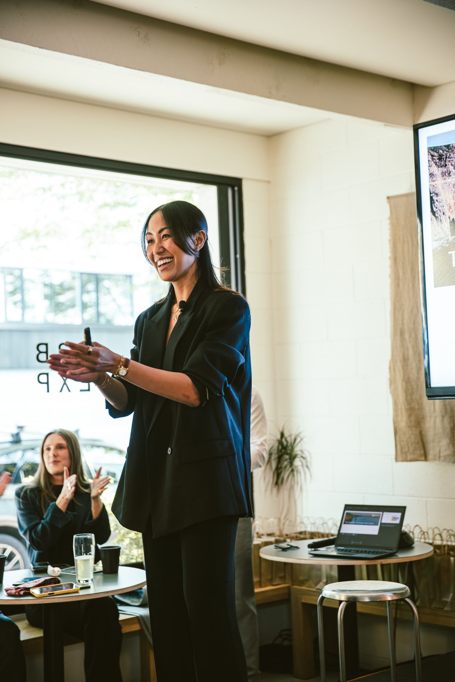 Smiling woman in black blazer presenting at a business event in a modern cafe setting.