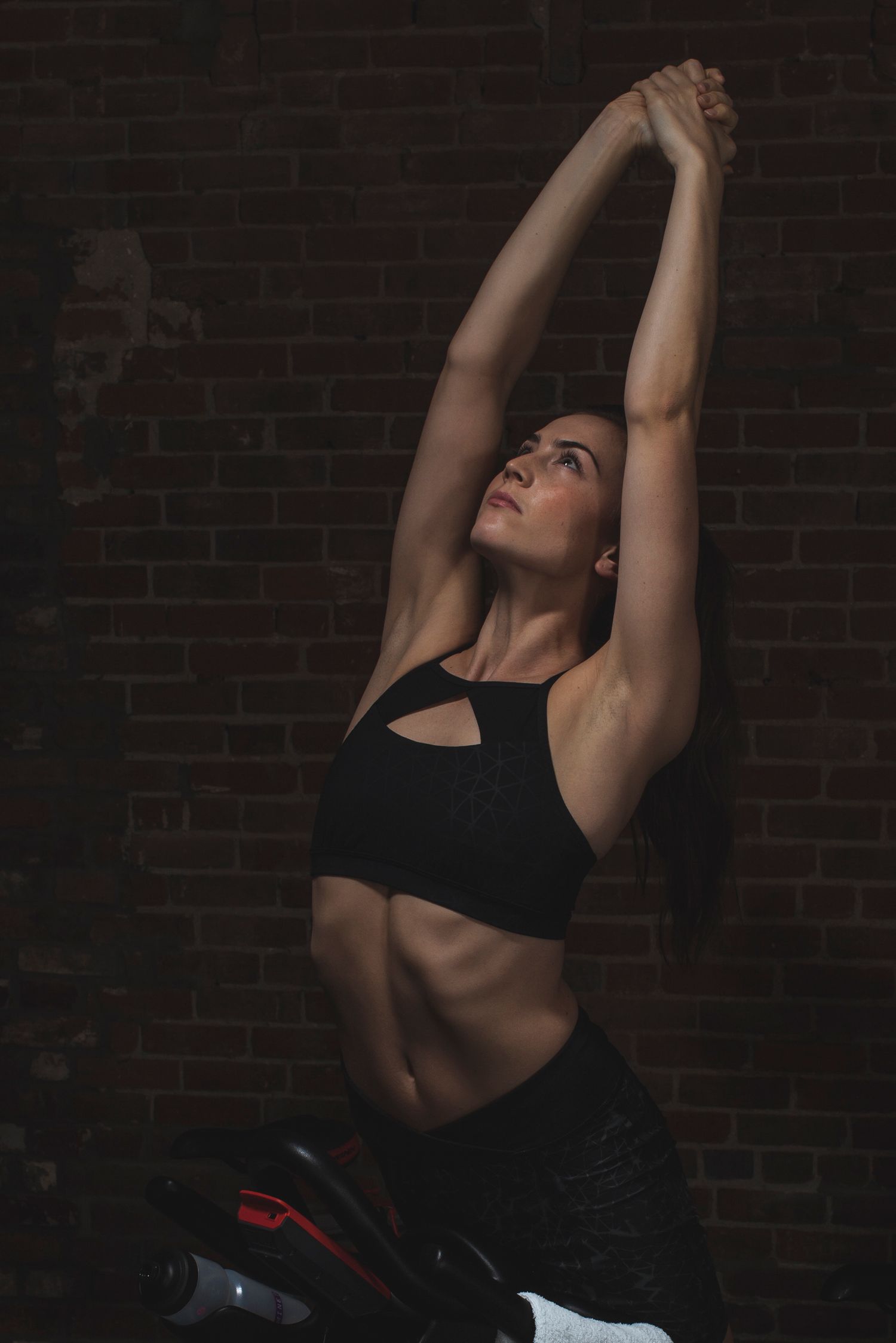 Fit woman in black sports bra stretching arms overhead against dark brick wall in gym workout pose.
