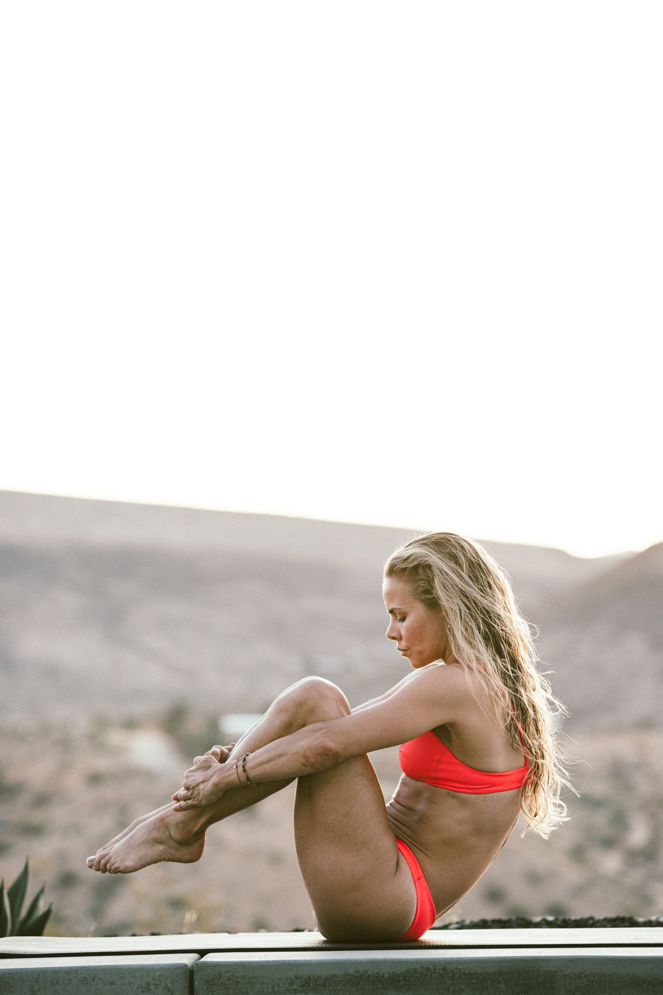 Fit blonde woman in red bikini sits on an outdoor ledge with desert mountains in the background.