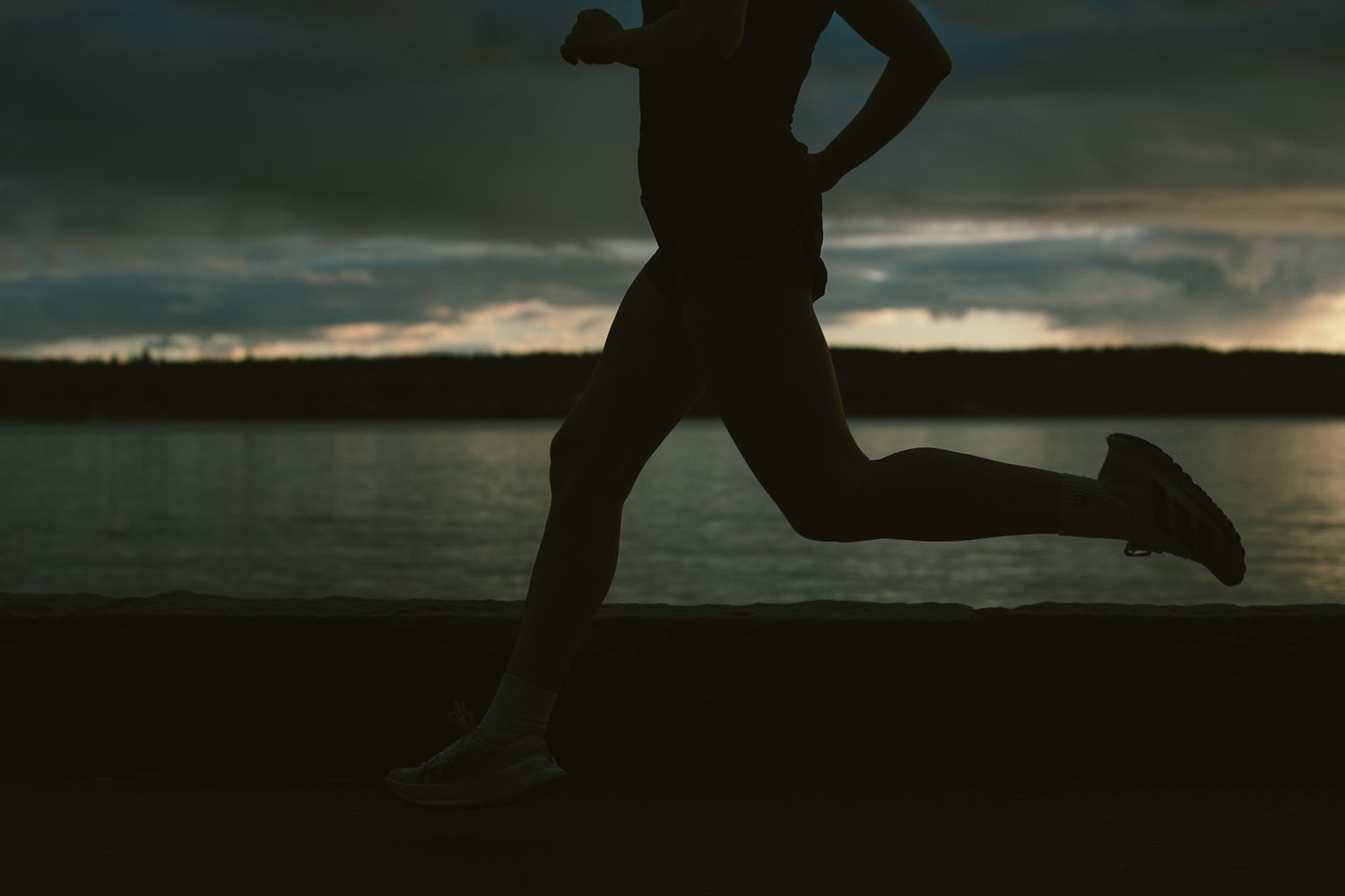 Silhouette of a runner in shorts and sneakers jogging along a waterfront wall at dramatic sunset.