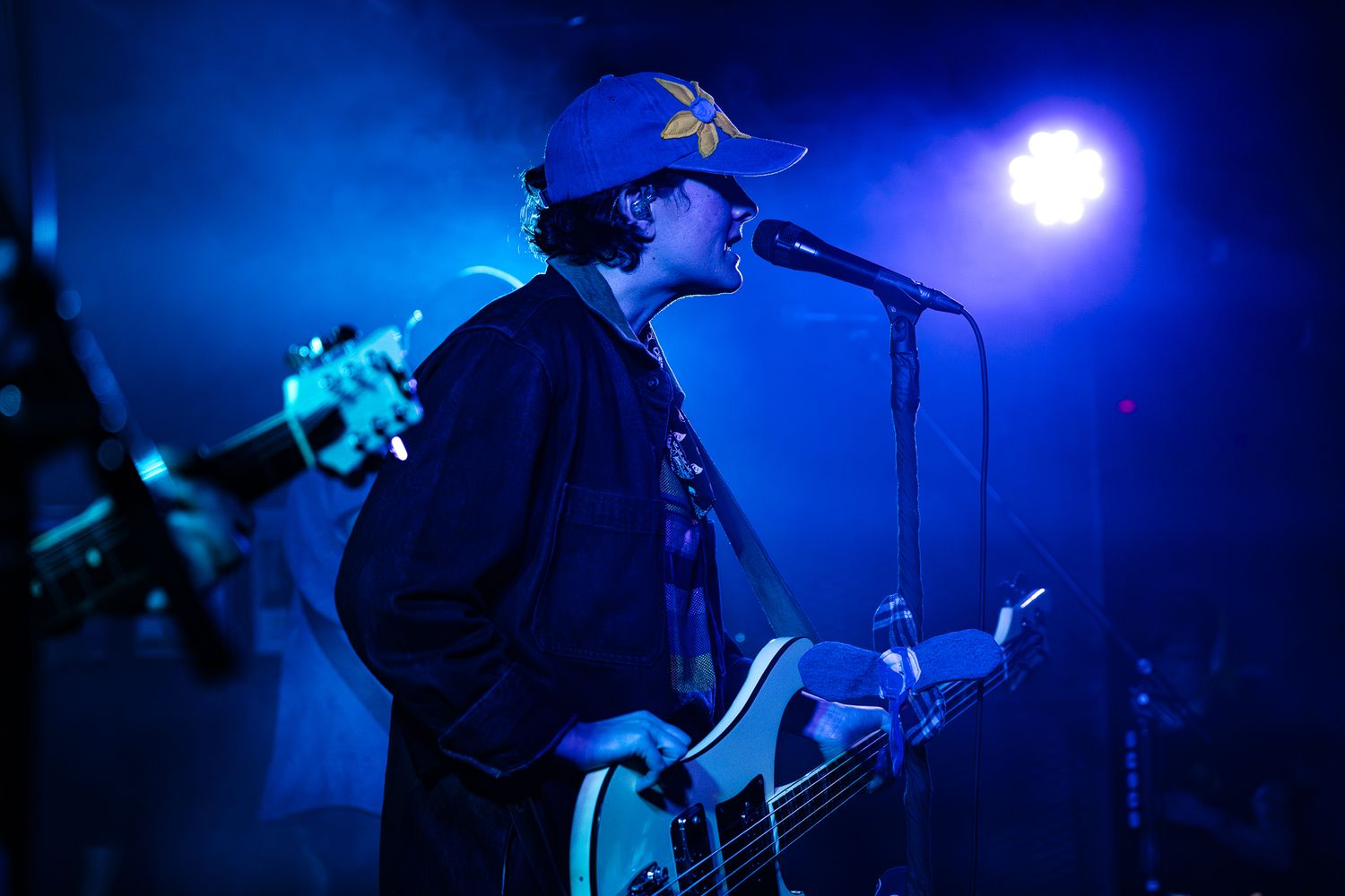 Guitarist in a blue cap performs on stage bathed in dramatic blue concert lighting while singing into a mic.