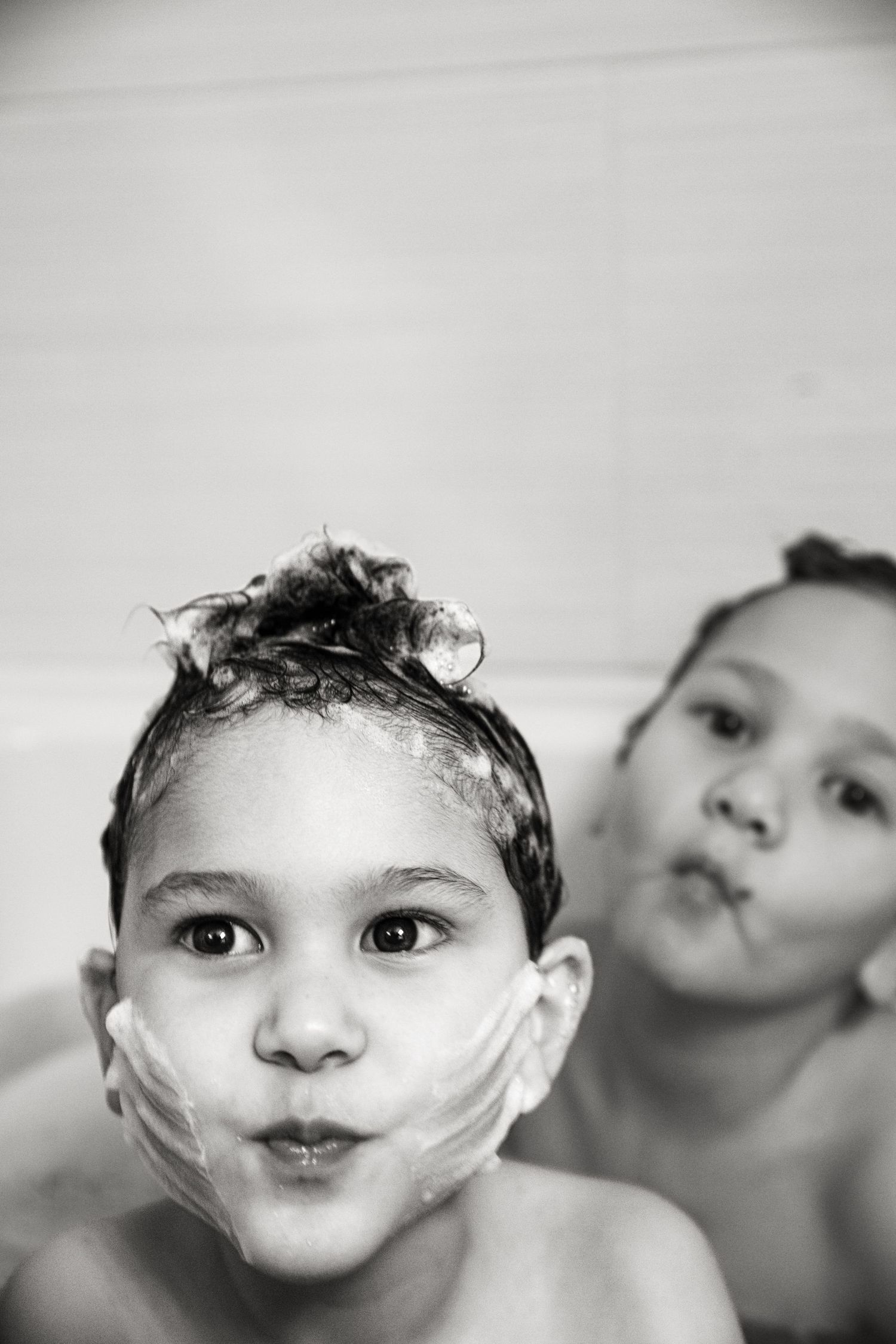 Black and white portrait of two young children with soapy hair making funny faces during bath time.