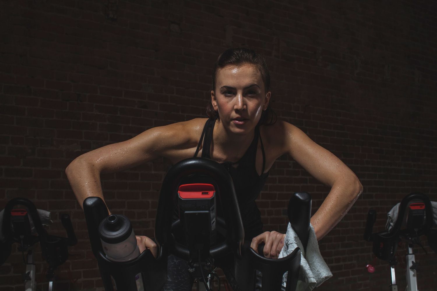 Determined woman in black sports bra cycling intensely on stationary bike in dark gym with brick wall.