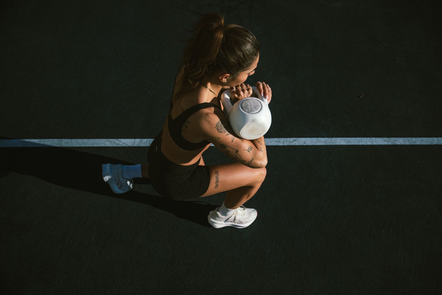 Athletic tattooed woman squatting low while holding a white kettlebell on a dark outdoor surface.