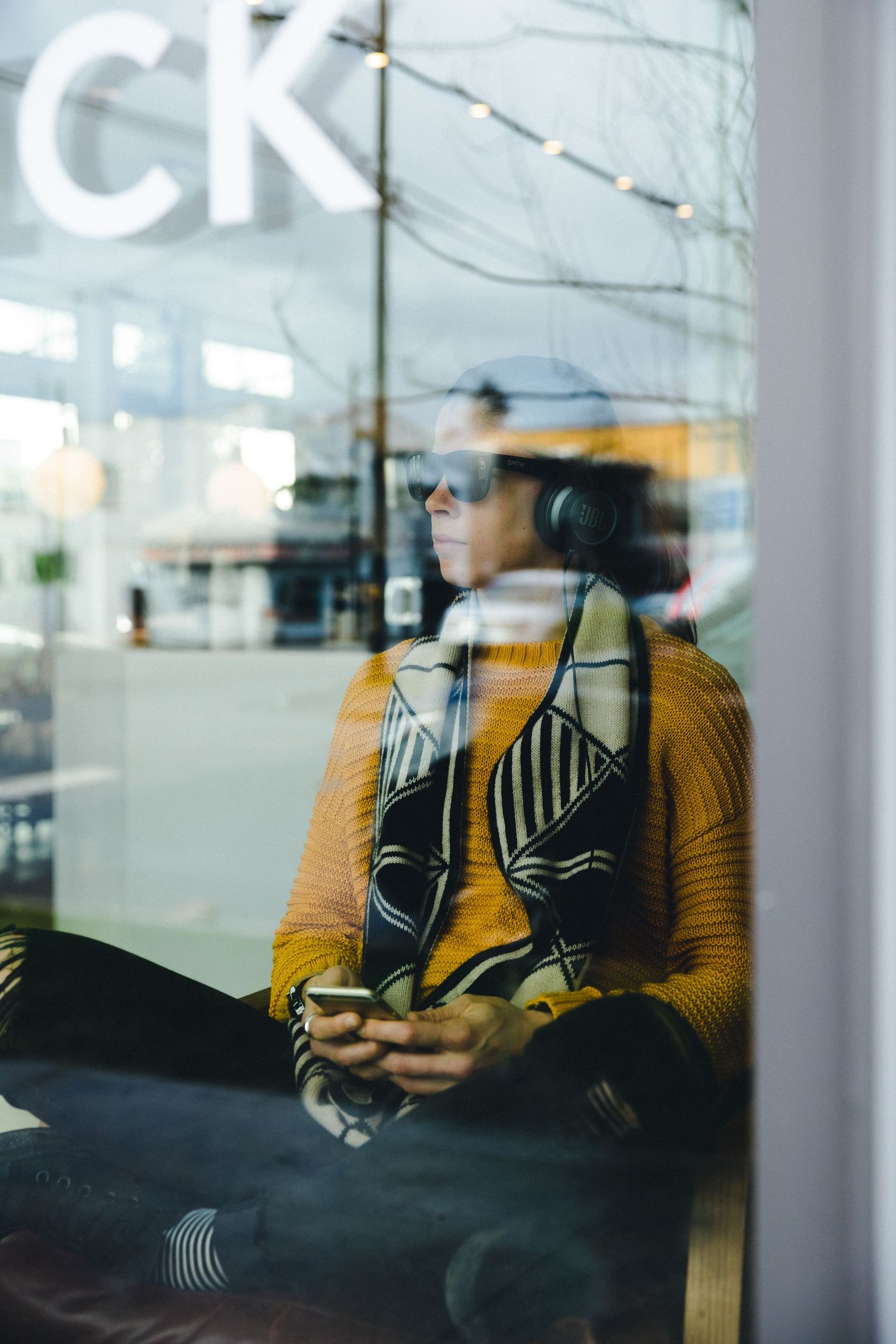 Person in yellow sweater with headphones and phone sits by a window, wearing sunglasses and a patterned scarf.