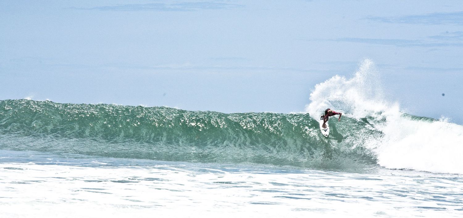 Surfer riding a large ocean wave with white water spray on a sunny day at the beach.