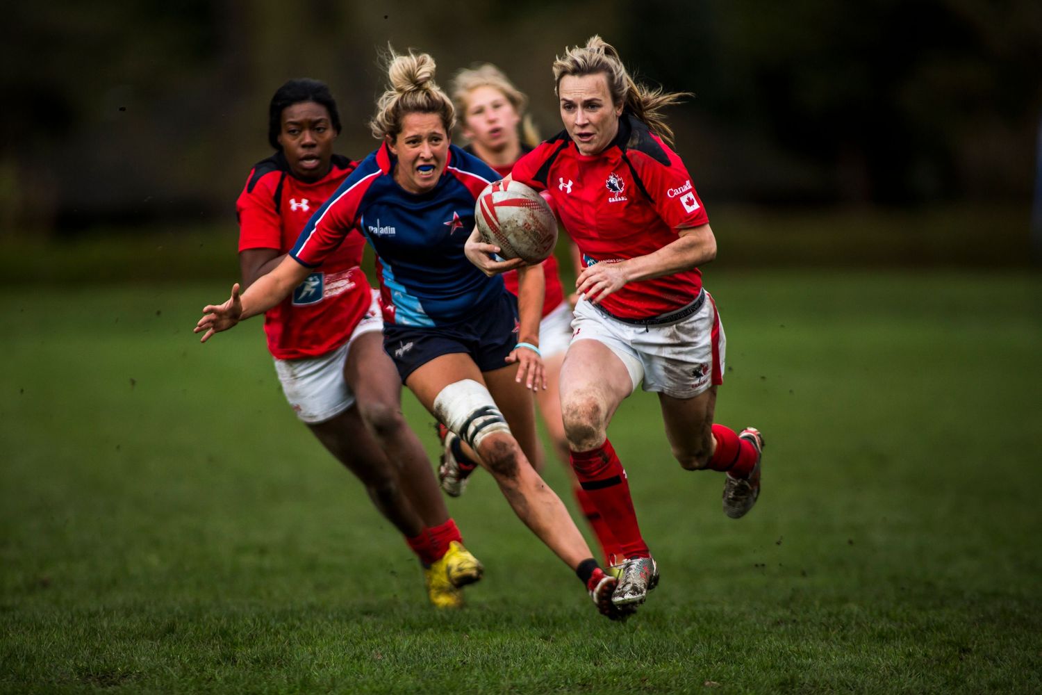 Women rugby players in red and blue uniforms compete intensely on a grass field during a match.