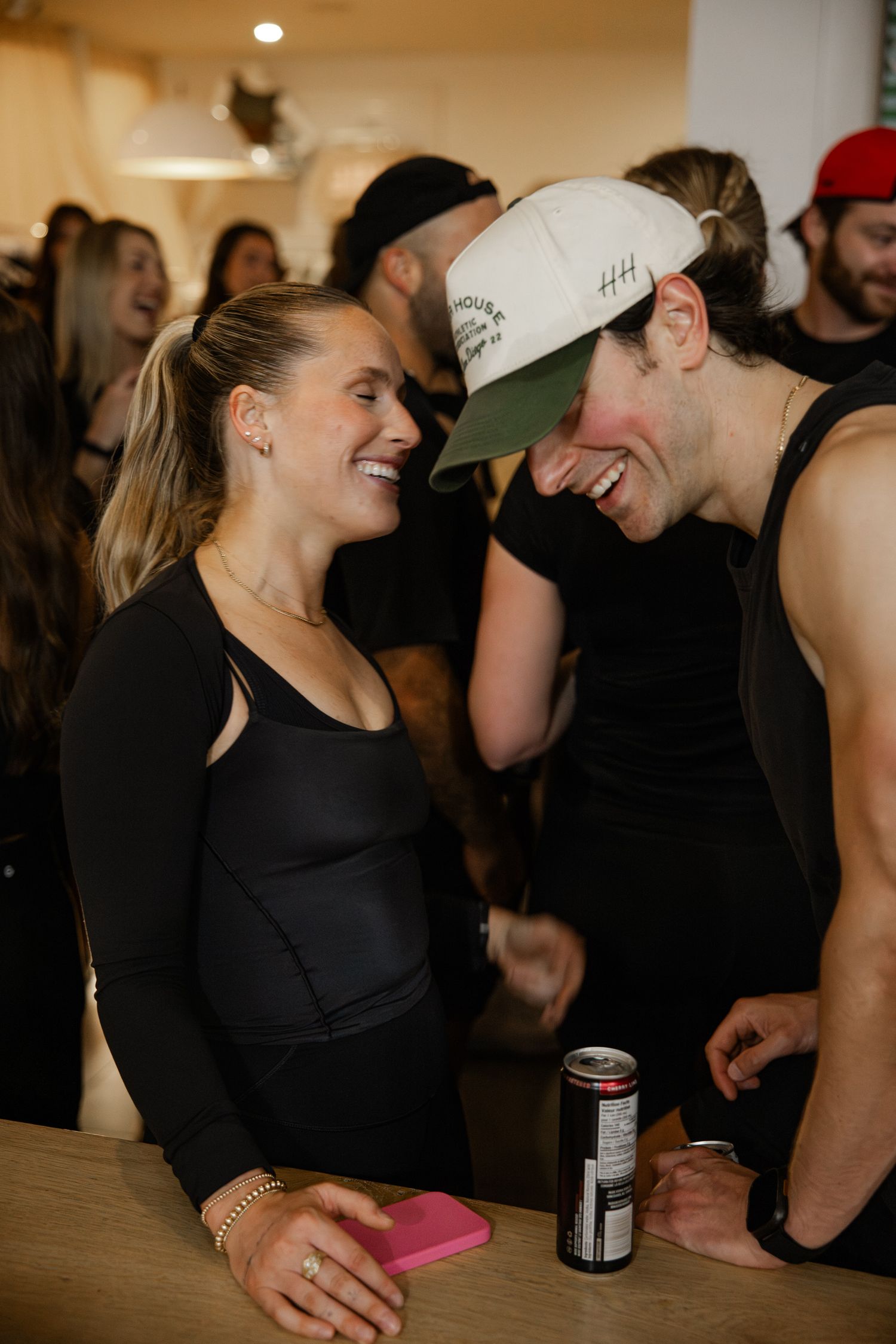 Two athletes laugh together at a post-race social event, one holding a pink phone and energy drink.