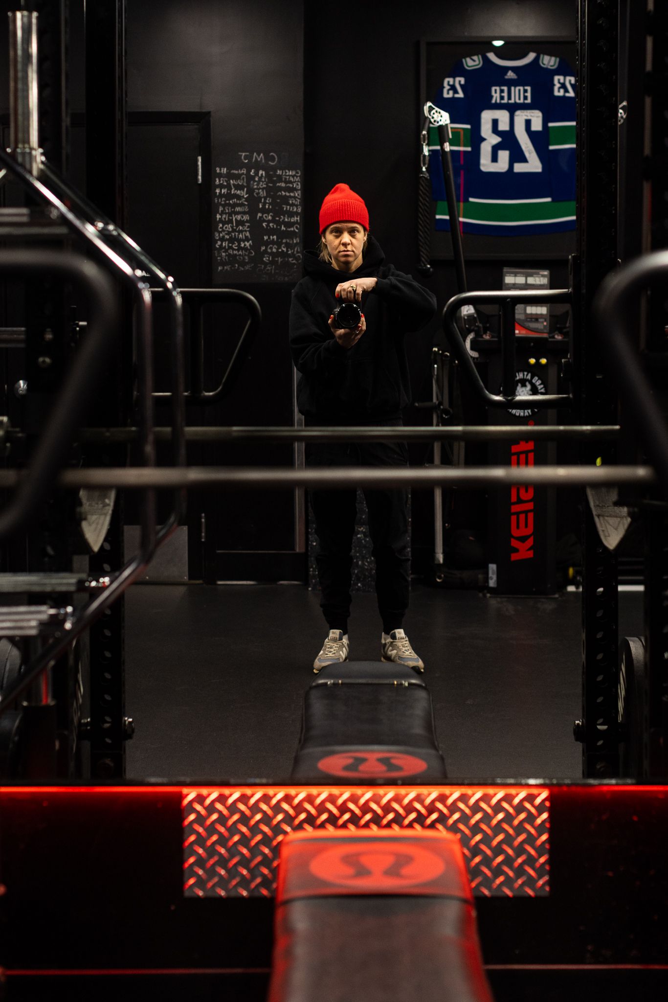 Man in red beanie standing in dark gym with hockey jersey displayed on wall behind him.