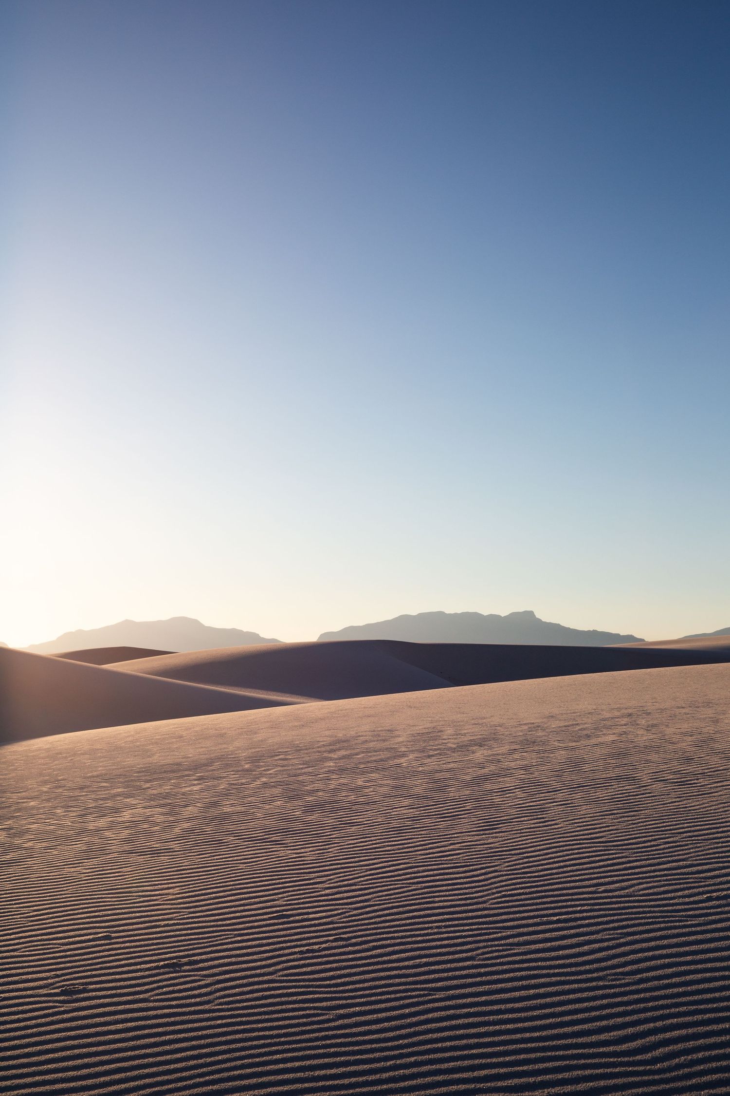 Serene white sand dunes at sunset with rippled texture, mountain silhouettes, and clear blue sky in a desert landscape.