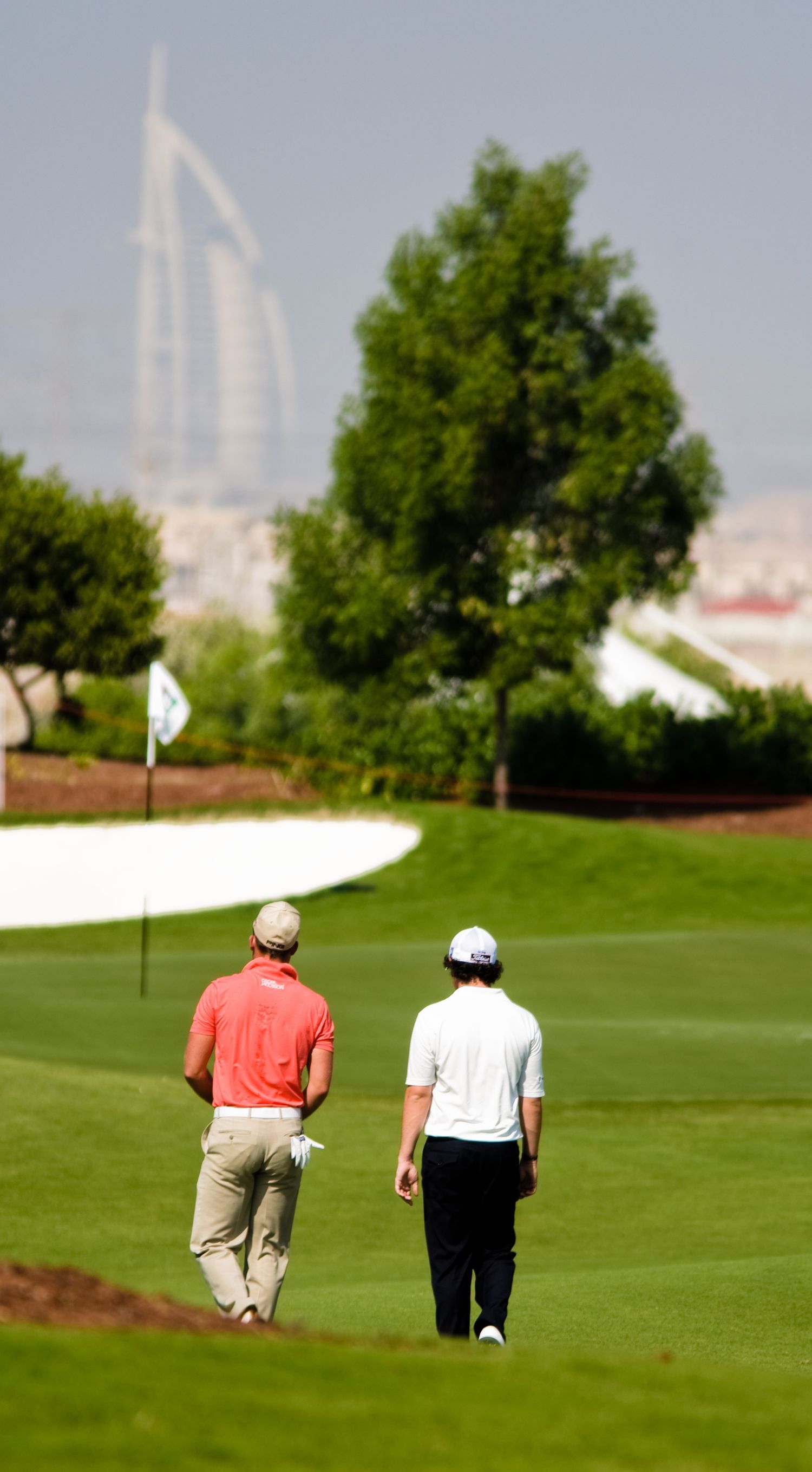 Two golfers walking on a lush green golf course with the iconic Burj Al Arab hotel visible in the background in Dubai.