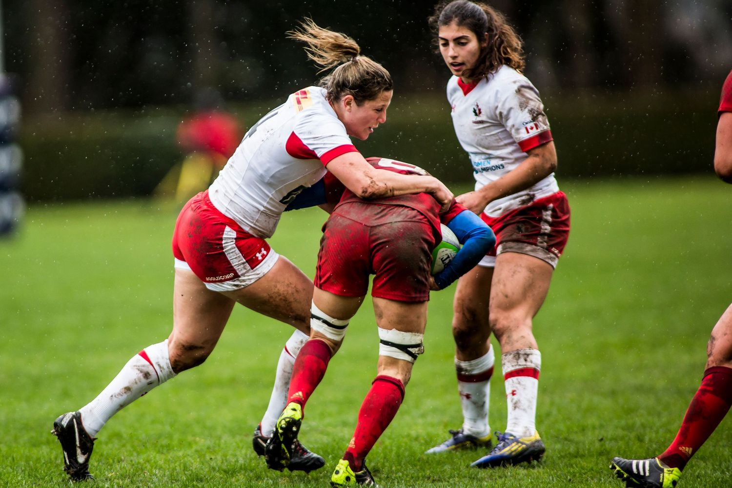 Women rugby players compete in a intense tackle during a rainy match, wearing red and white jerseys.
