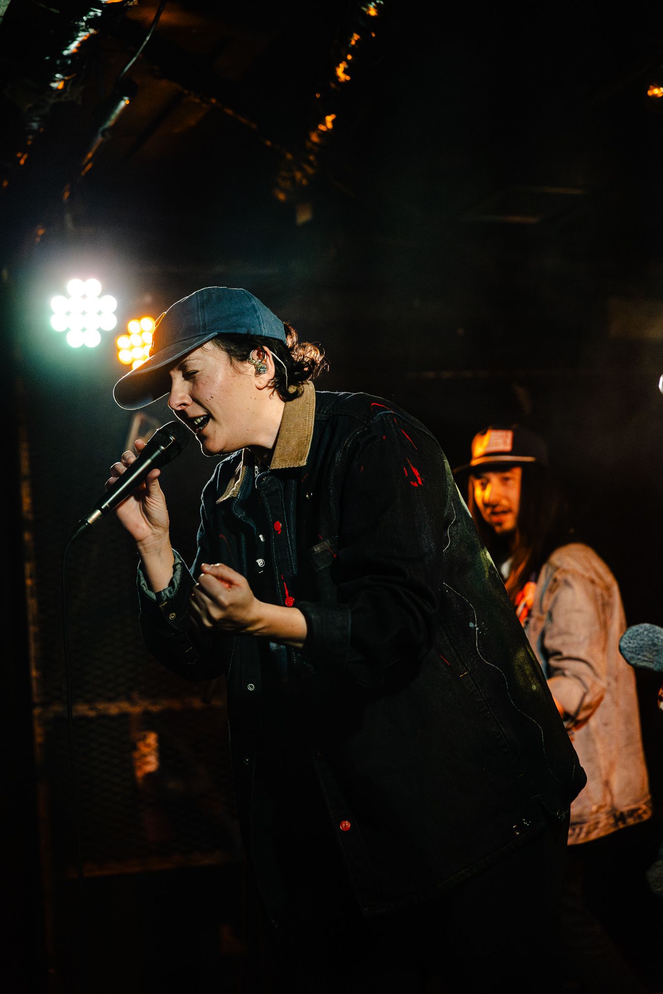 Young performer in a snapback cap sings passionately into a microphone on a dimly lit concert stage.