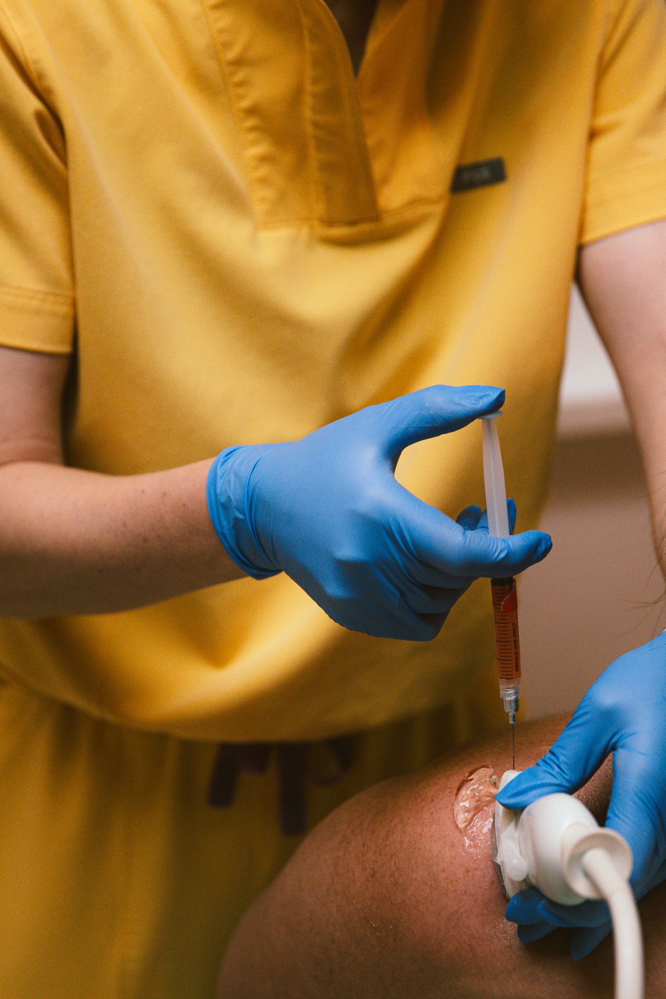 Medical professional in yellow scrubs and blue gloves administers an injection into a patient's knee joint.