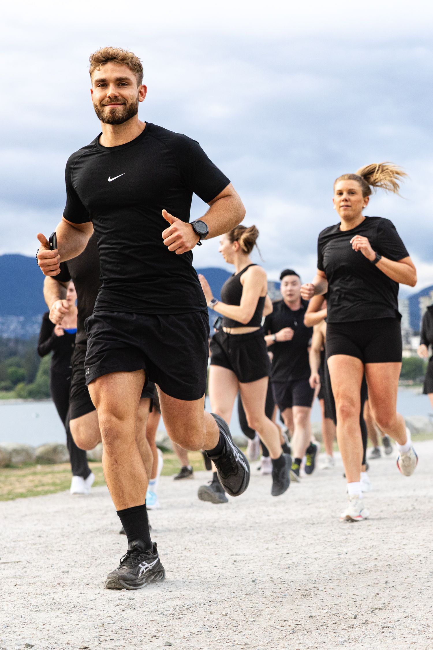 Smiling man in black Nike outfit leads a group of runners along a waterfront path outdoors.