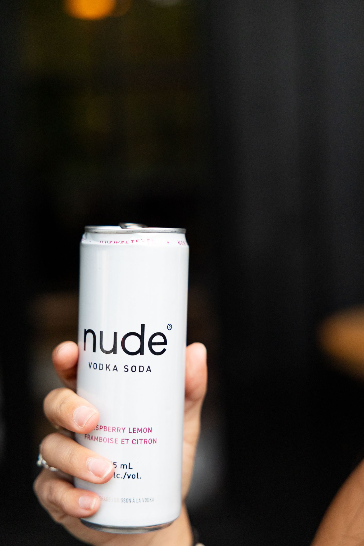 Hand holding a Nude Vodka Soda raspberry lemon flavored can against a dark background.