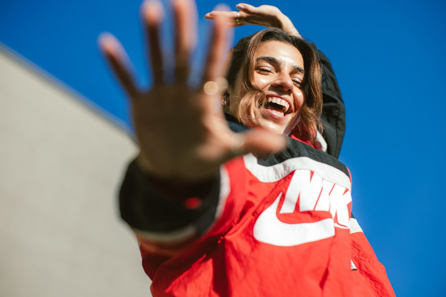 Joyful young person in a red Nike windbreaker reaching hand toward camera under a blue sky.